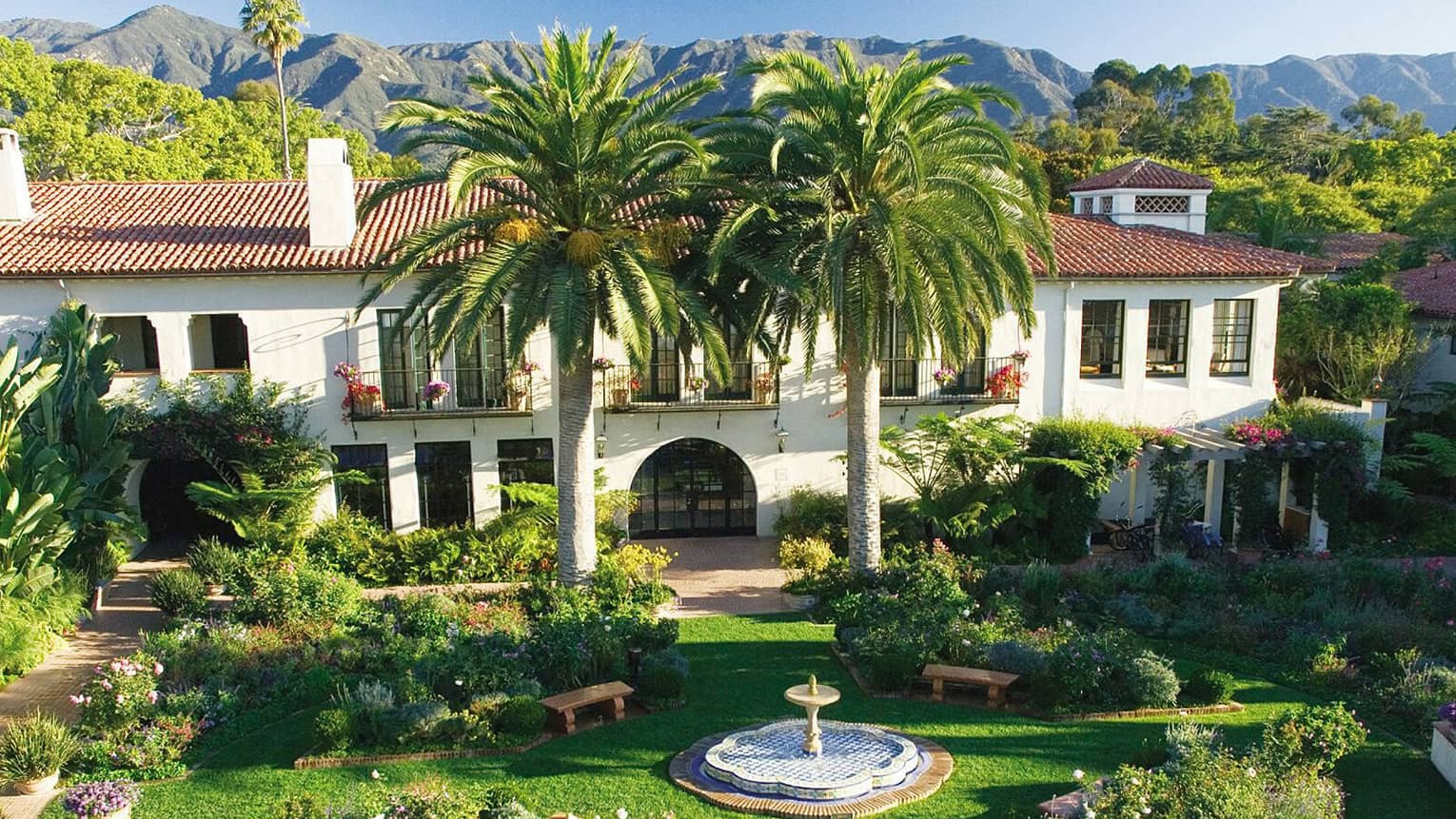 Lush green courtyard with a fountain in the center, flanked by two large palm trees and a Spanish Colonial-style building in the background, overlooking the mountains