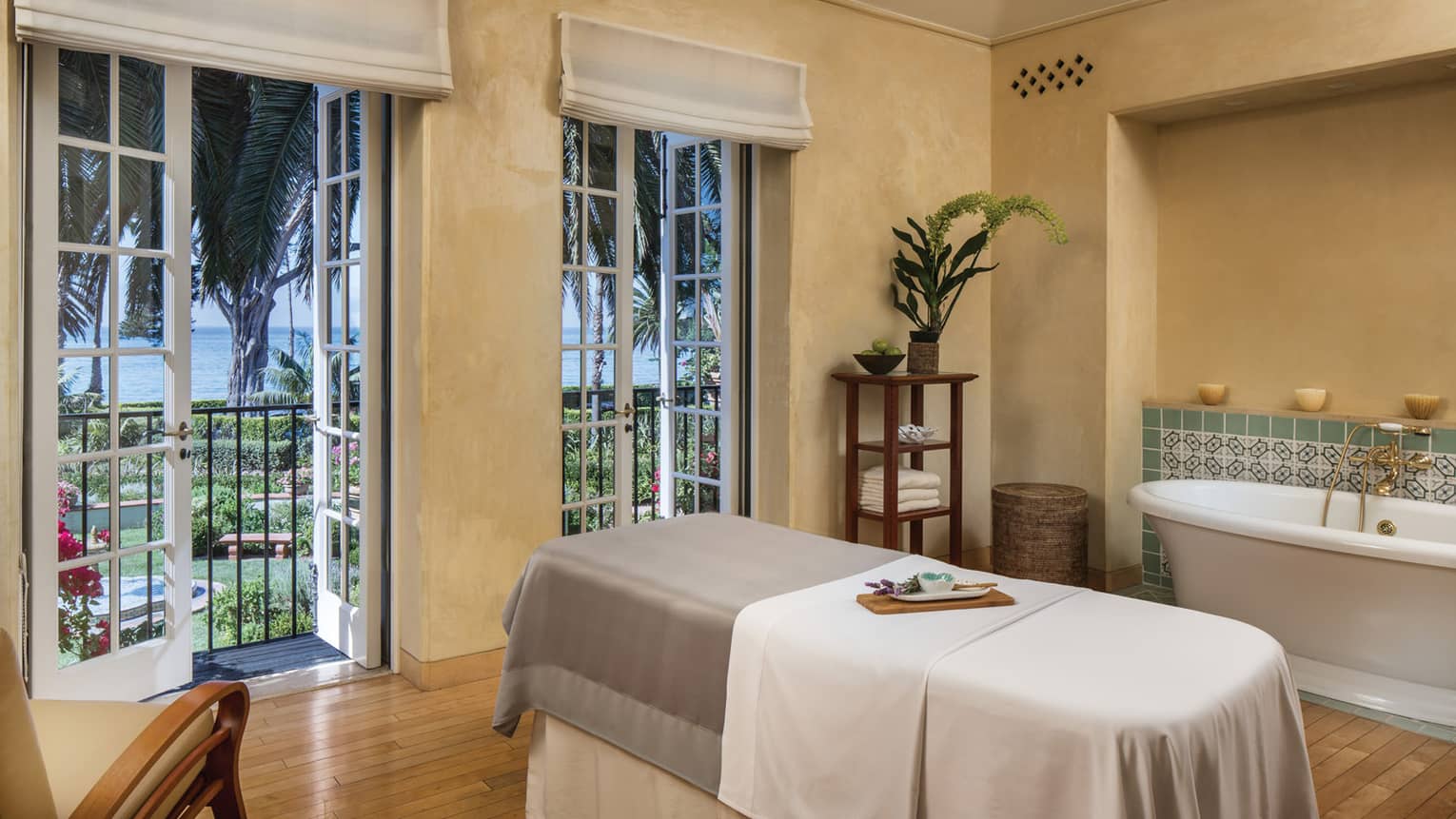 A spa treatment room with massage table, bath tub, wooden floors, French doors to a garden