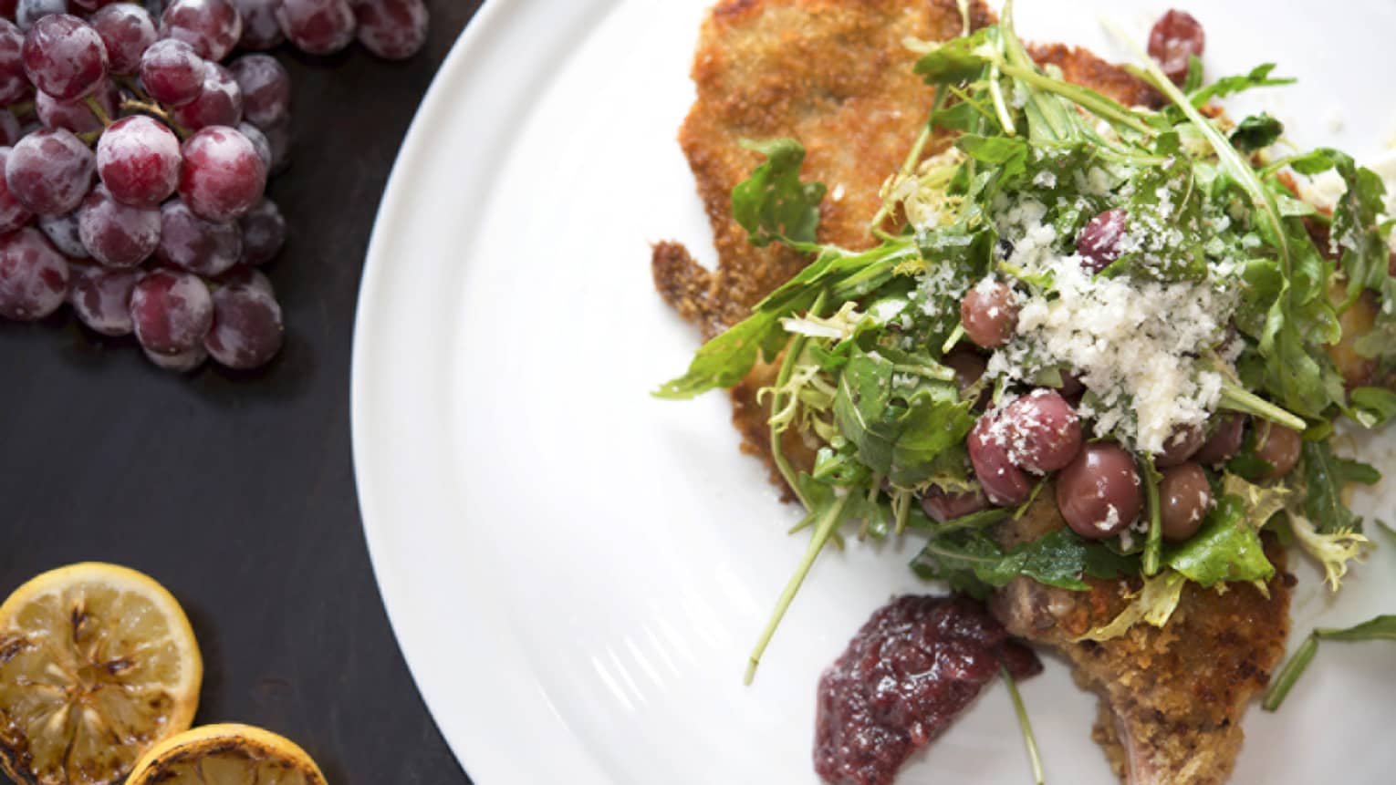 A white porcelain plate with a fired piece of fish covered with arugula, olives, feta cheese with grapes and lemons on the side