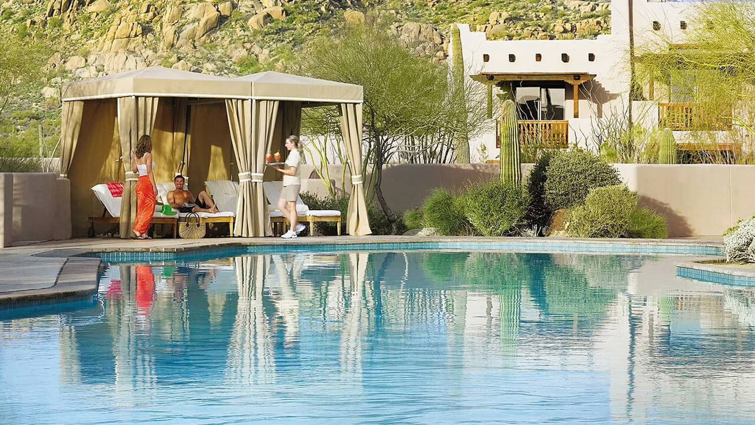 Server brings tray of cocktails to woman, man under pool cabana on sunny swimming pool deck