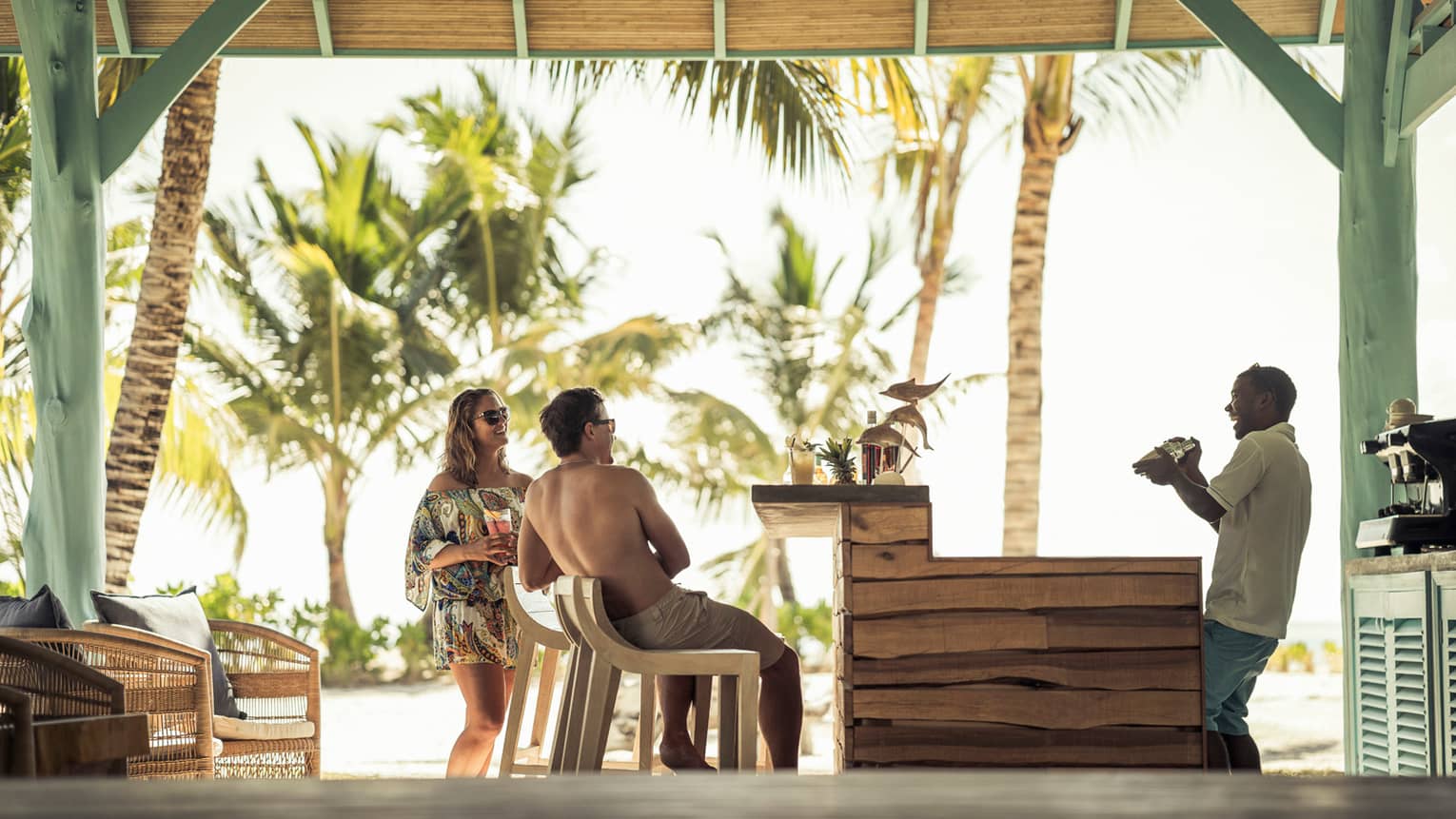 Couple in beach wear laugh with bartender who shakes cocktail in thatched roof bar
