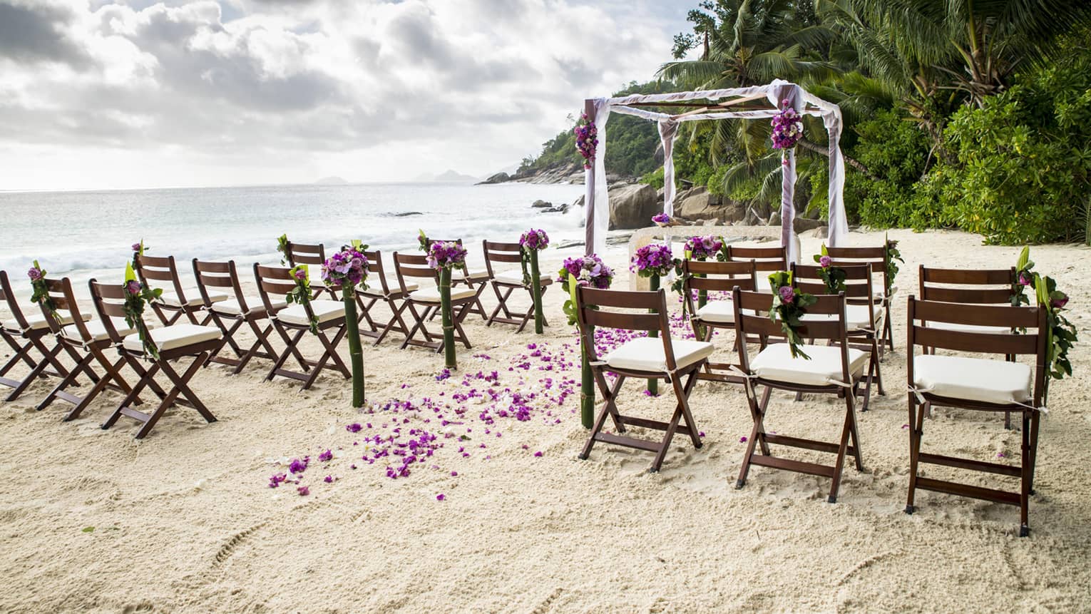 Rows of wooden chairs on sand in front of canopy decorated with purple flowers by ocean