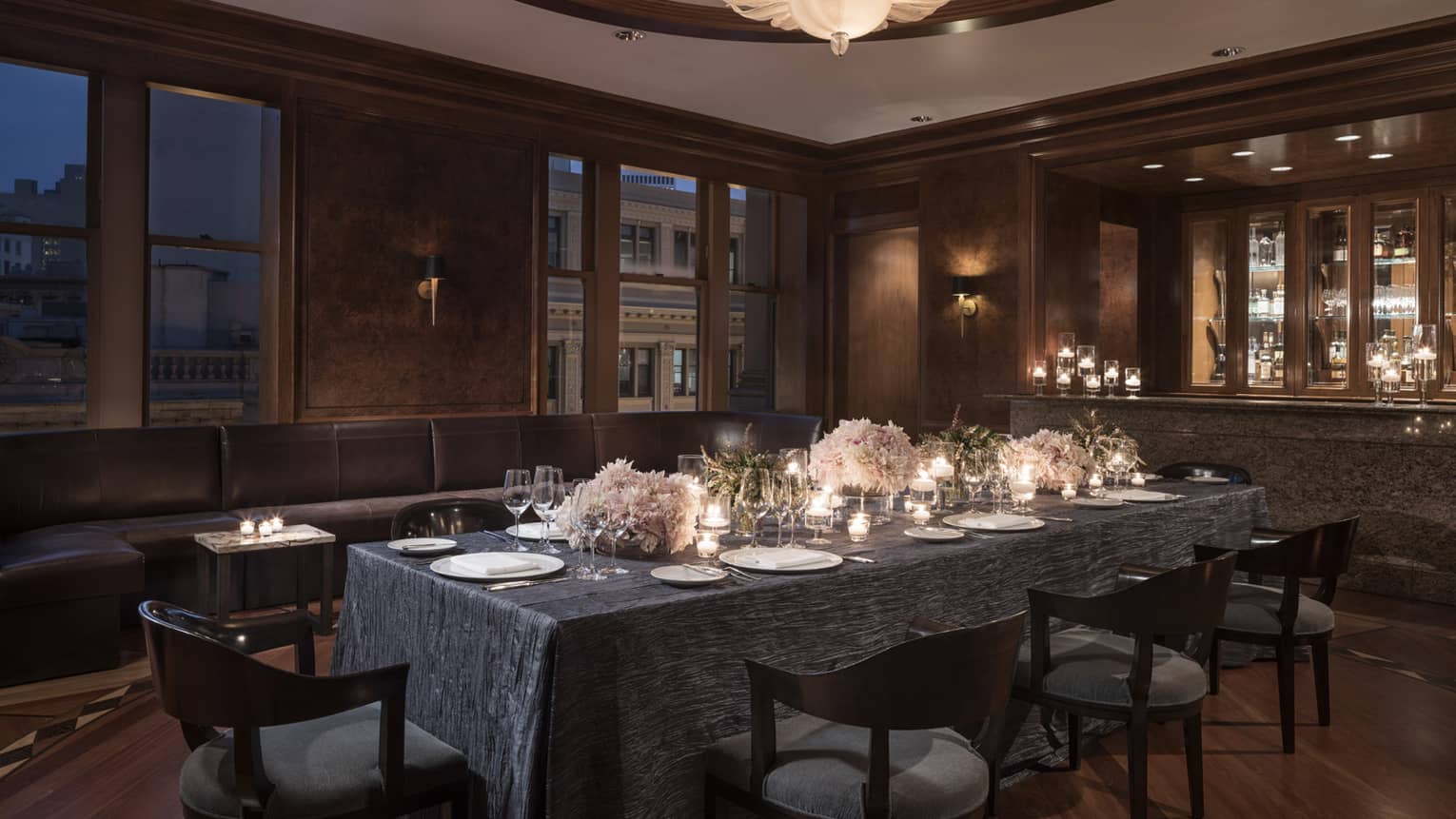 Private dining table with candles, flowers near long leather banquette, windows at night