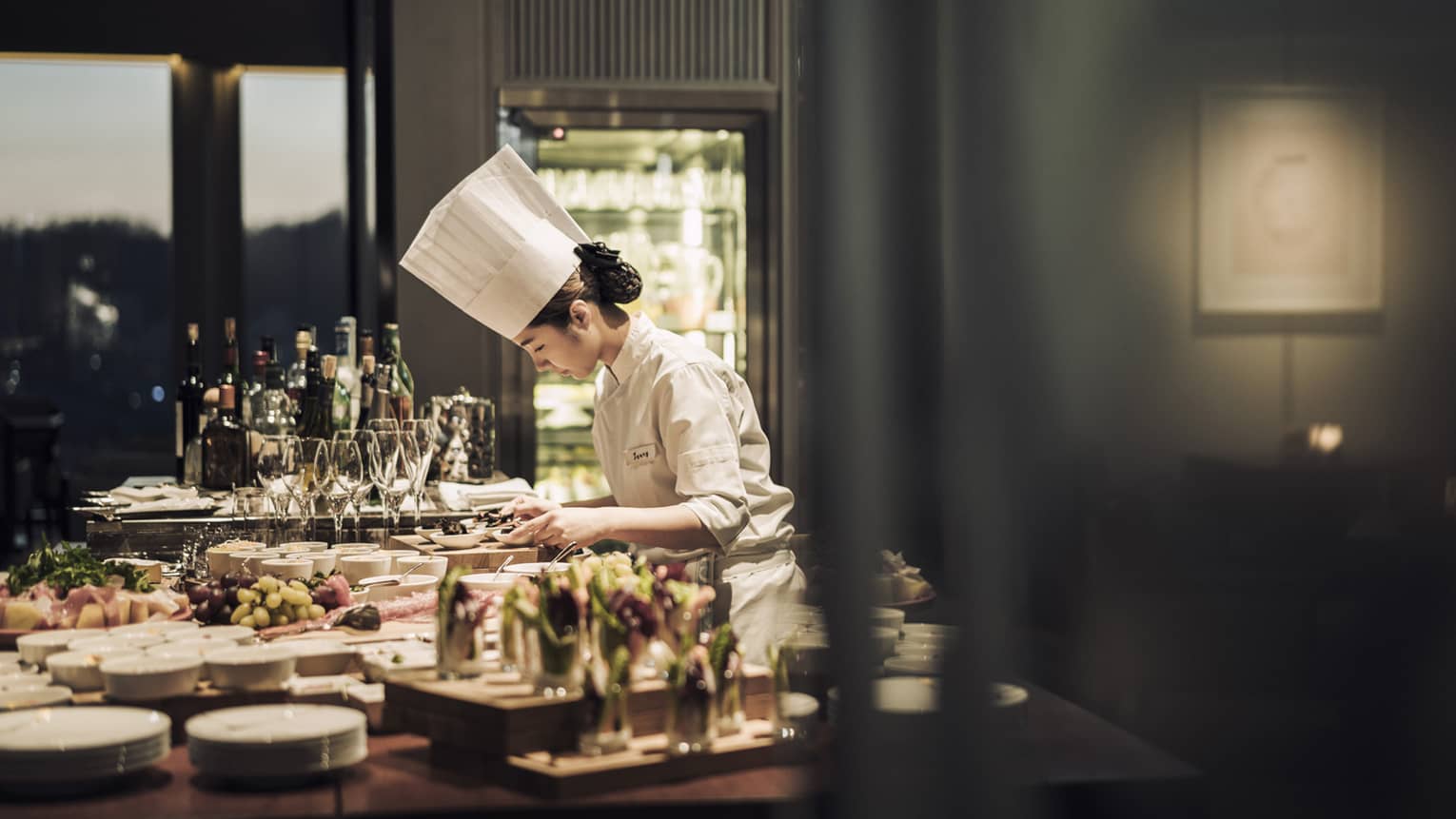 Chef wearing white uniform, hat stands garnishes appetizers on counter covered with dishes, meals
