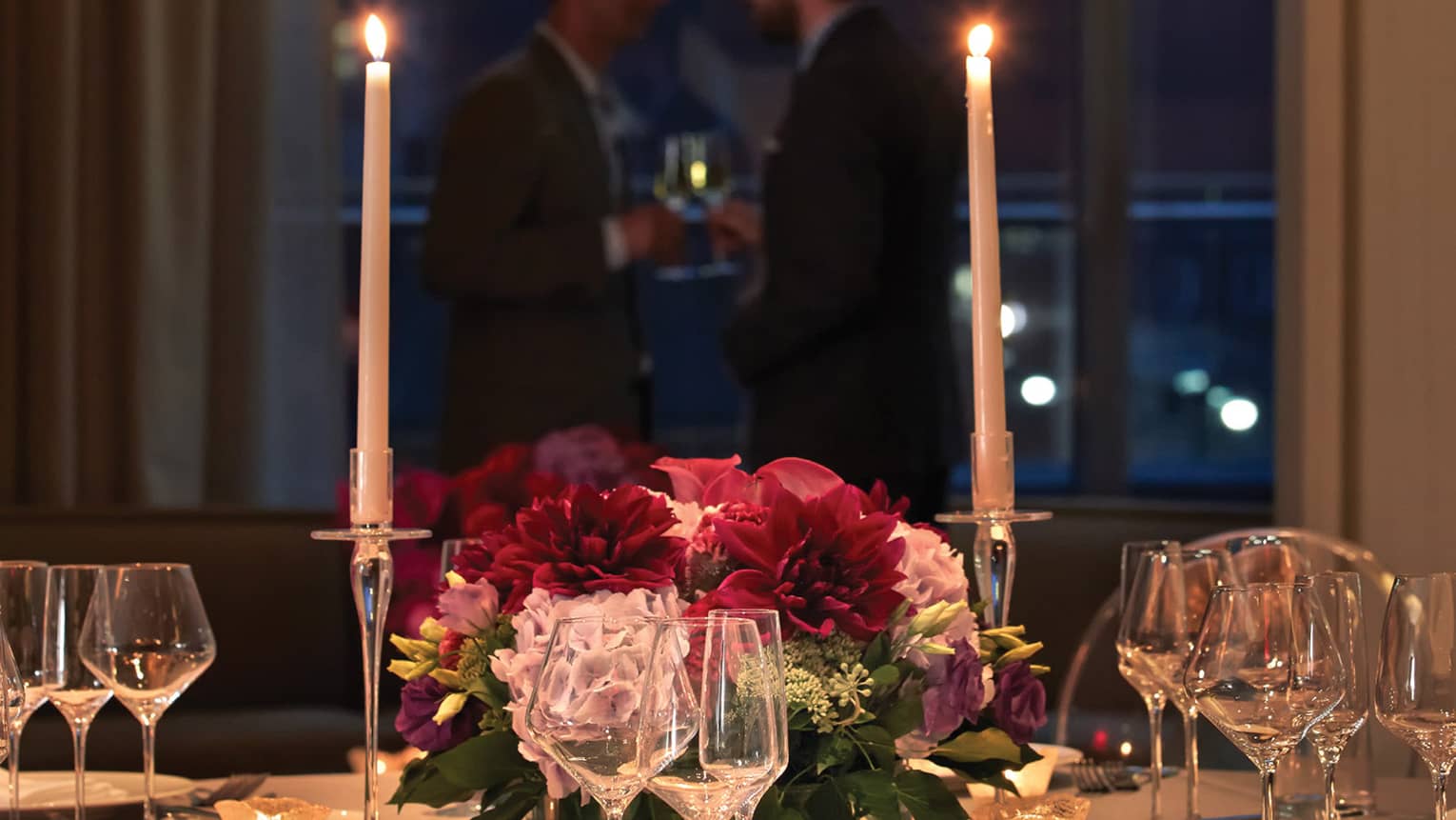 Two tall glowing candles and floral arrangement, glassware on dining table