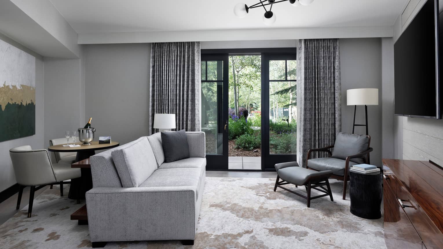 Guest living room with grey sofa, grey leather accent chair, small round dining table, glass doors opening onto terrace, area rug