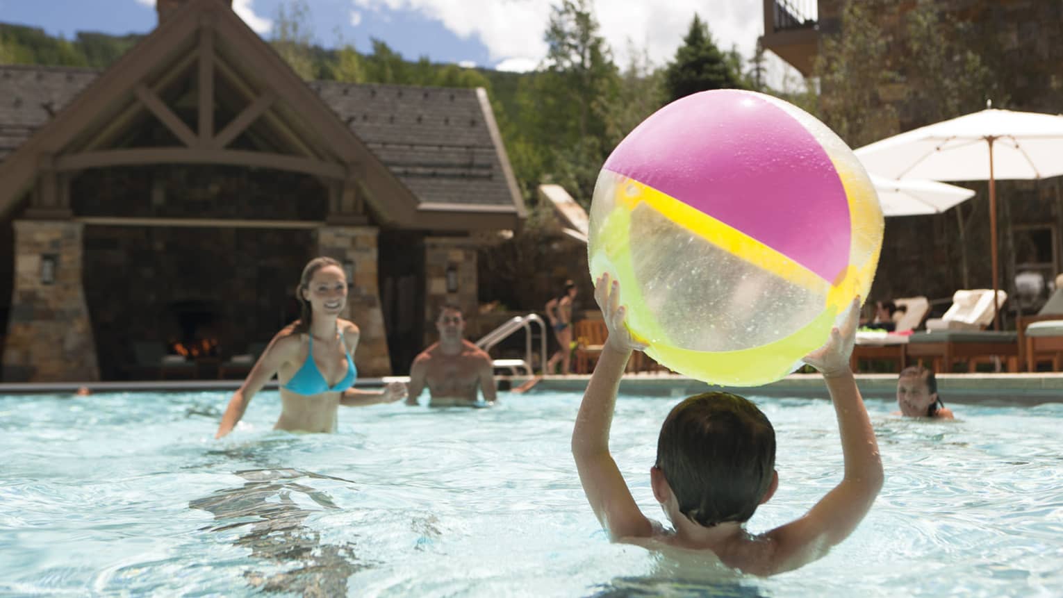 Child holds large, colorful beach ball over his head in swimming pool, prepares to throw it to parents nearby