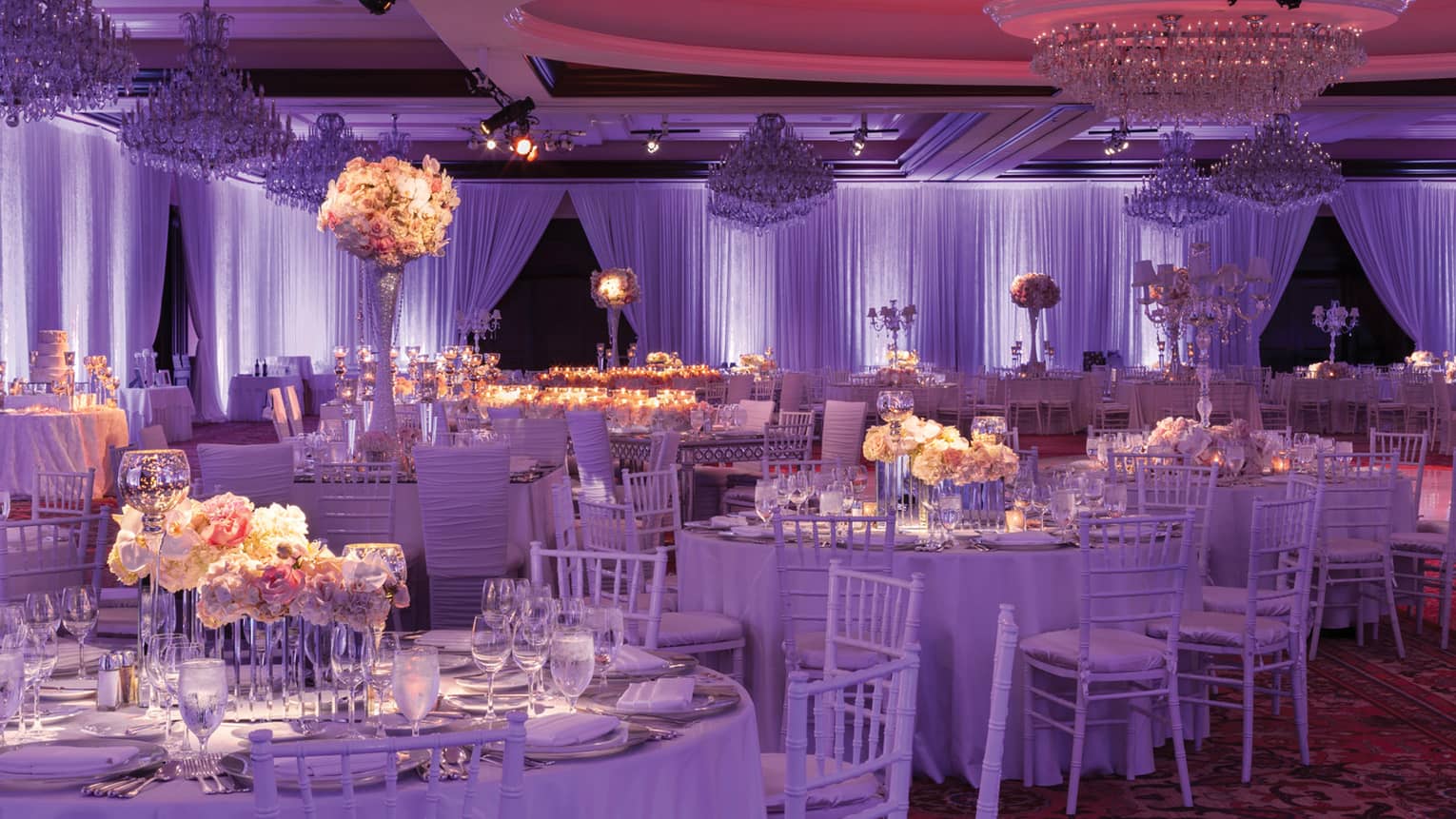 Wedding reception white tables and chairs, pink floral arrangements in ballroom with purple lights