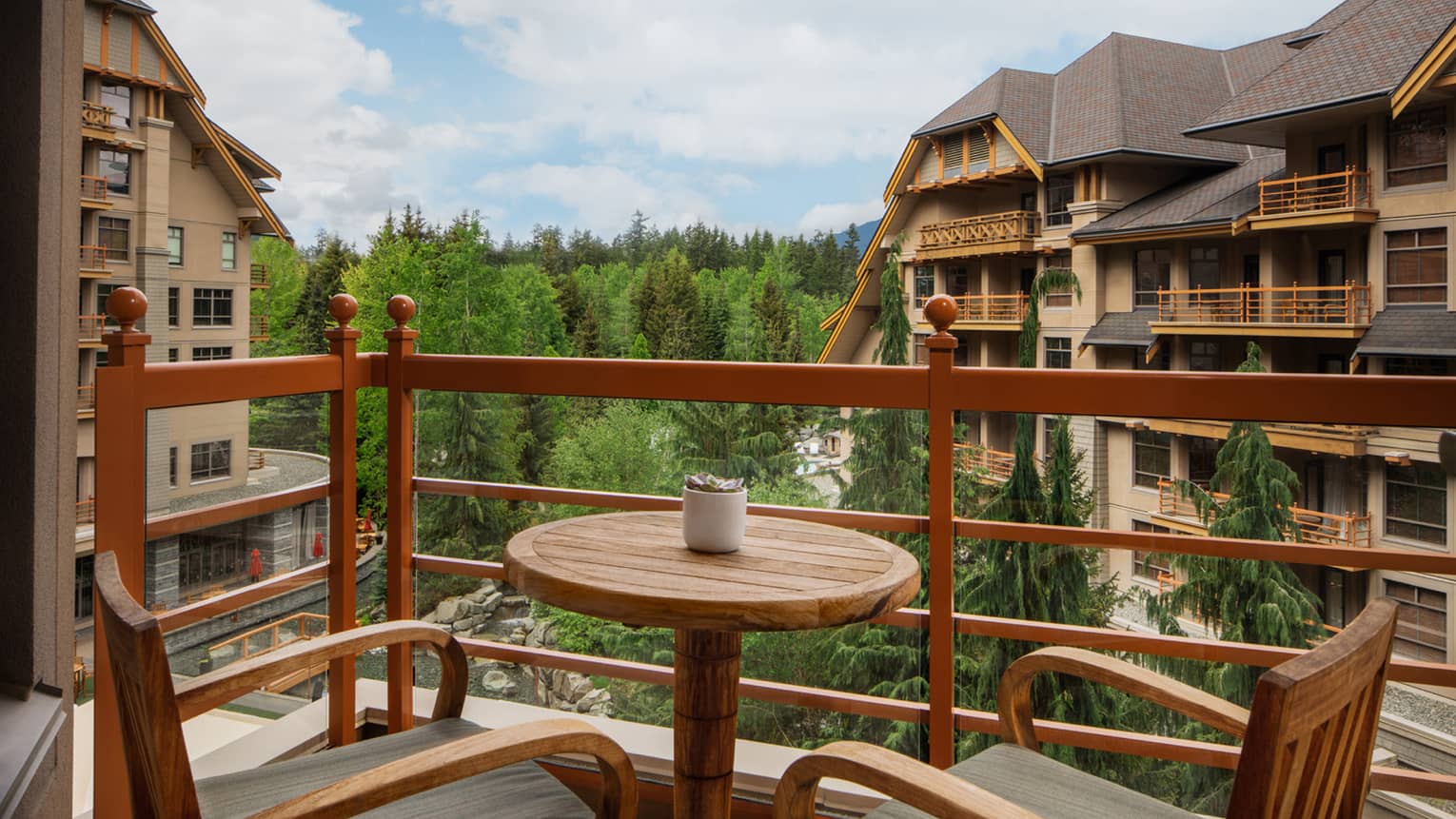 A picturesque view from a balcony of a Mountain View room at the Four Seasons Whistler. The balcony is furnished with wooden chairs and a round table, perfect for enjoying the outdoors. It overlooks other chalet-style buildings amid a lush green forest backdrop, highlighting the serene and natural setting of the resort.