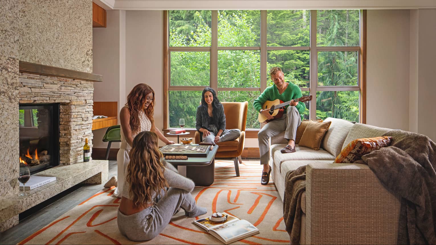 Cozy gathering in a luxury vacation rental living area at Four Seasons Whistler. Four people enjoy leisure time together; one man plays guitar while seated on a chair, and three women engage in conversation and board games. The room is warmly decorated with a stone fireplace, large windows showcasing a forest view, a plush sofa, and vibrant geometric-patterned rugs.