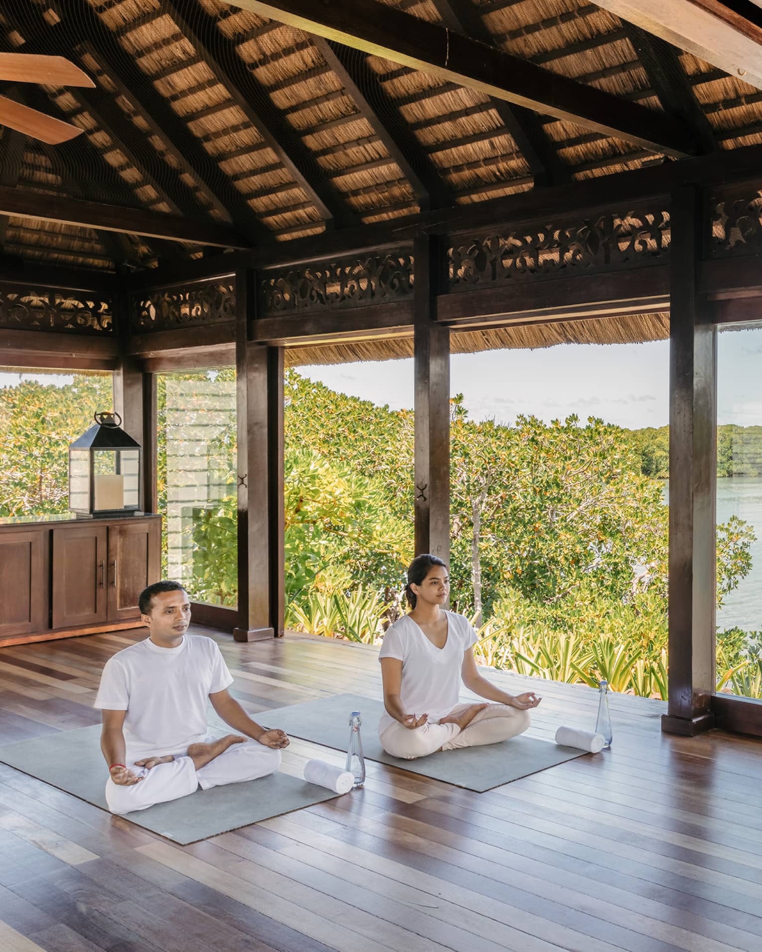 Two people wearing white seated in yoga poses in open-air spa yoga pavilion by beach