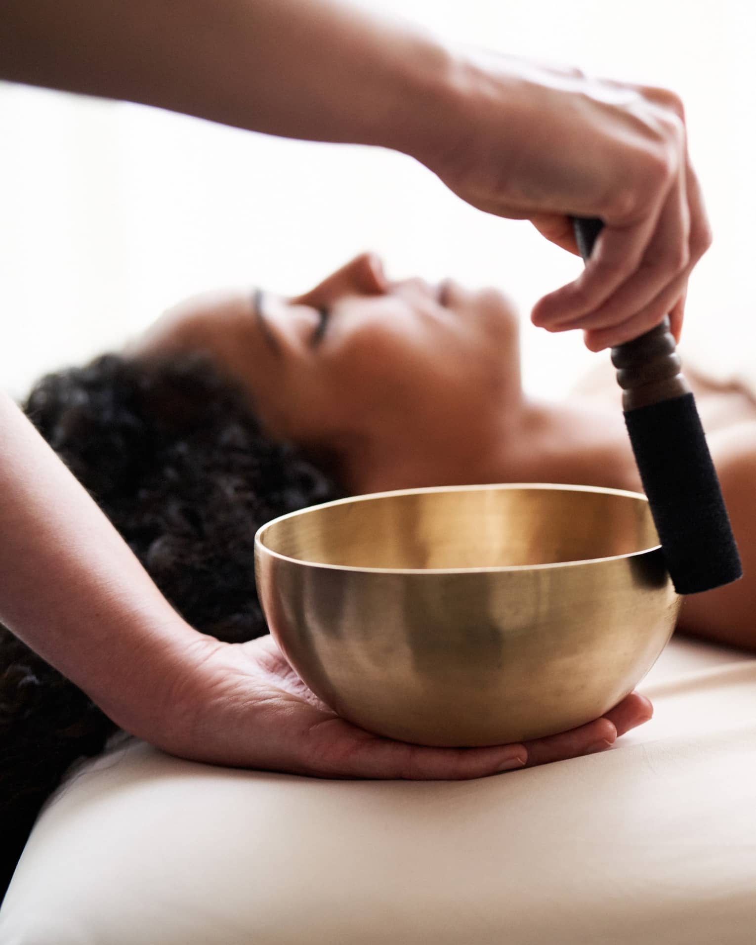 Woman with eyes closed lies on massage table in as vibrational sound therapy is performed with a Tibetan singing bowl