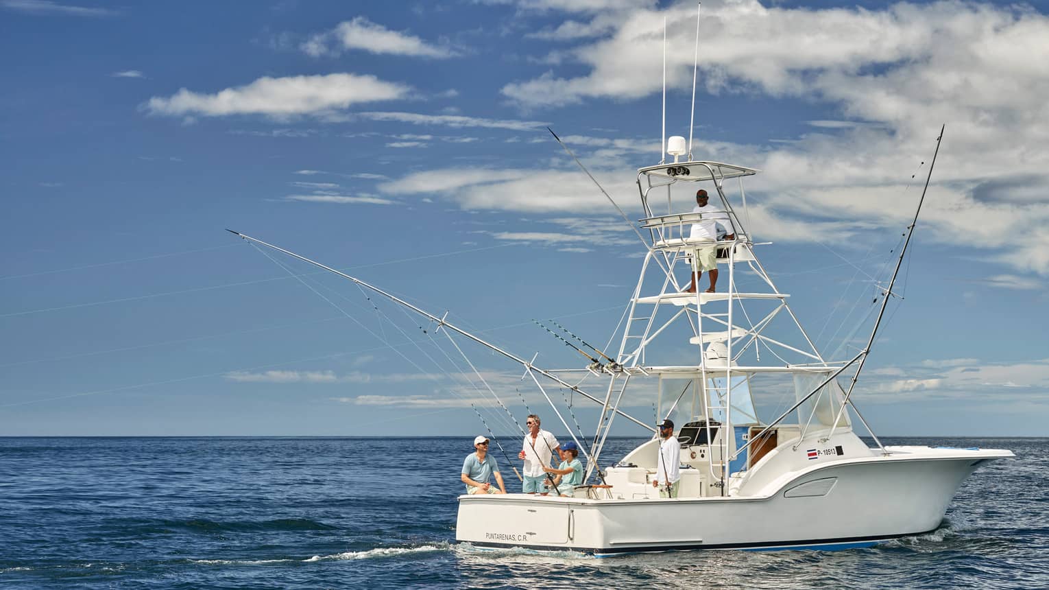 Four smiling guests on a fishing boat, a guide perched on a high platform lookout, on deep-blue water under a clear sky.