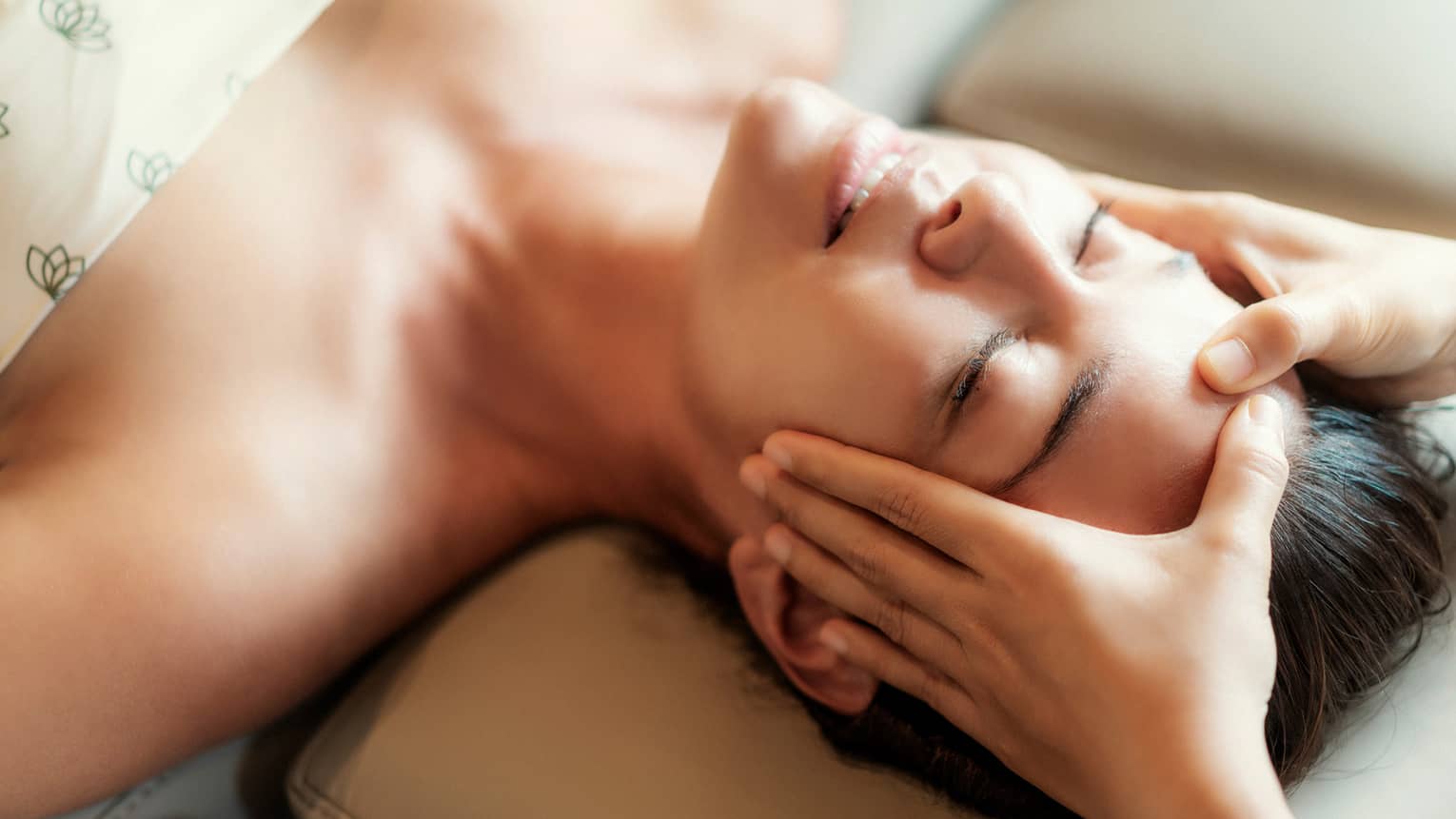 Spa facial, two hands rest on woman's forehead as she closes her eyes, lays under sheet on treatment table