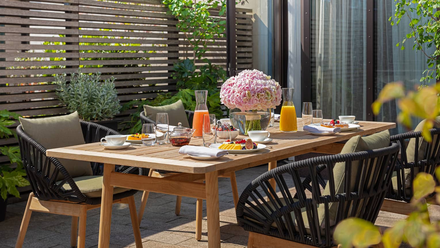 The Executive Conservatory at Four Seasons London Park Lane in Mayfair features an inviting outdoor dining area. A rustic wooden table is set with a colorful breakfast spread, including fresh fruits, pastries, juices, and tea. The table is beautifully decorated with a large bouquet of pink hydrangeas, enhancing the fresh, vibrant setting. Stylish black wicker chairs with comfortable cushions surround the table, nestled amidst lush greenery and a modern wooden slatted privacy screen. The scene is sunlit, offering a pleasant and serene dining experience.