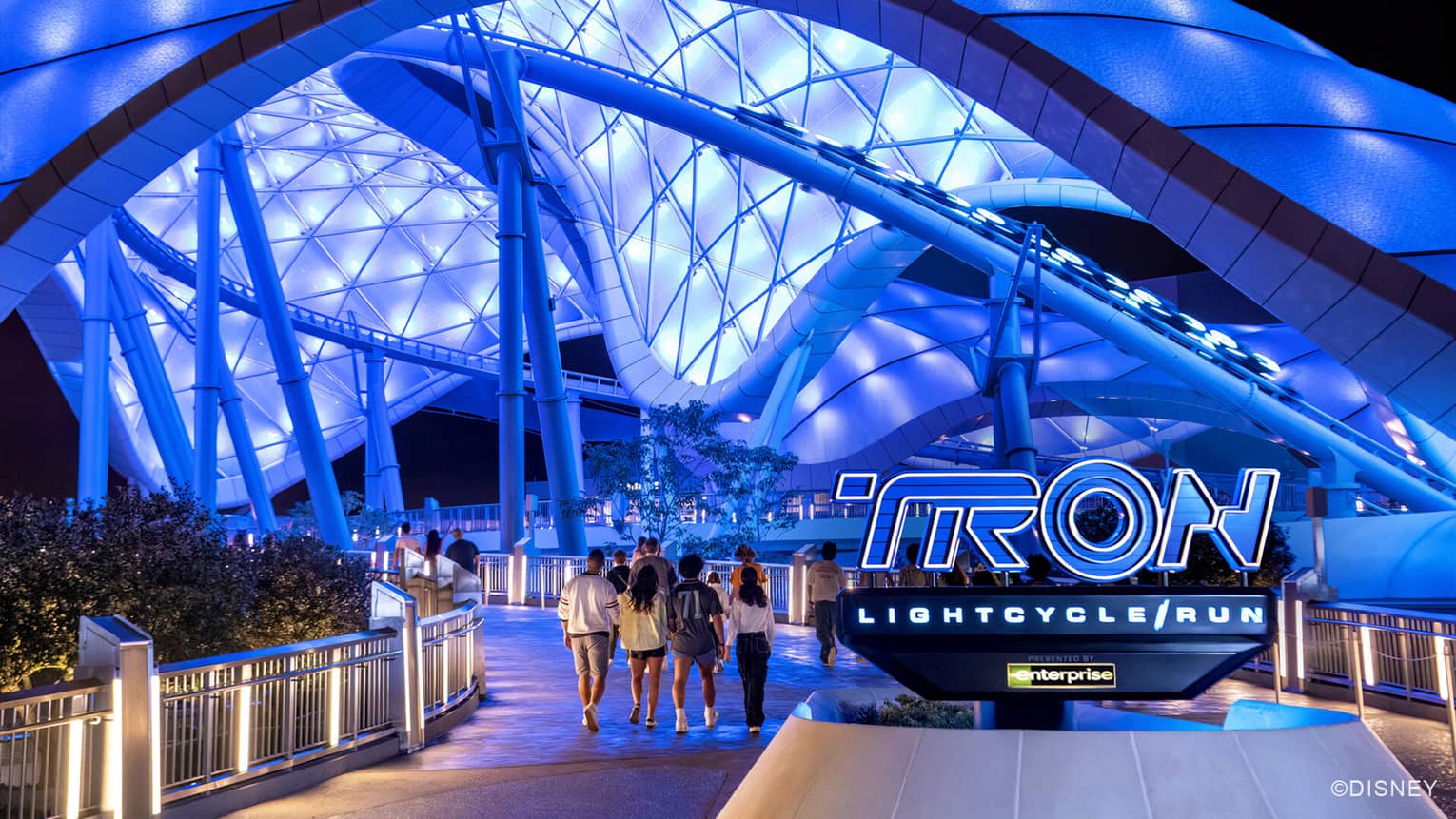 People walking under a glowing blue structure with a sign that says "TRON LIGHTCYCLE/RUN" under it.
