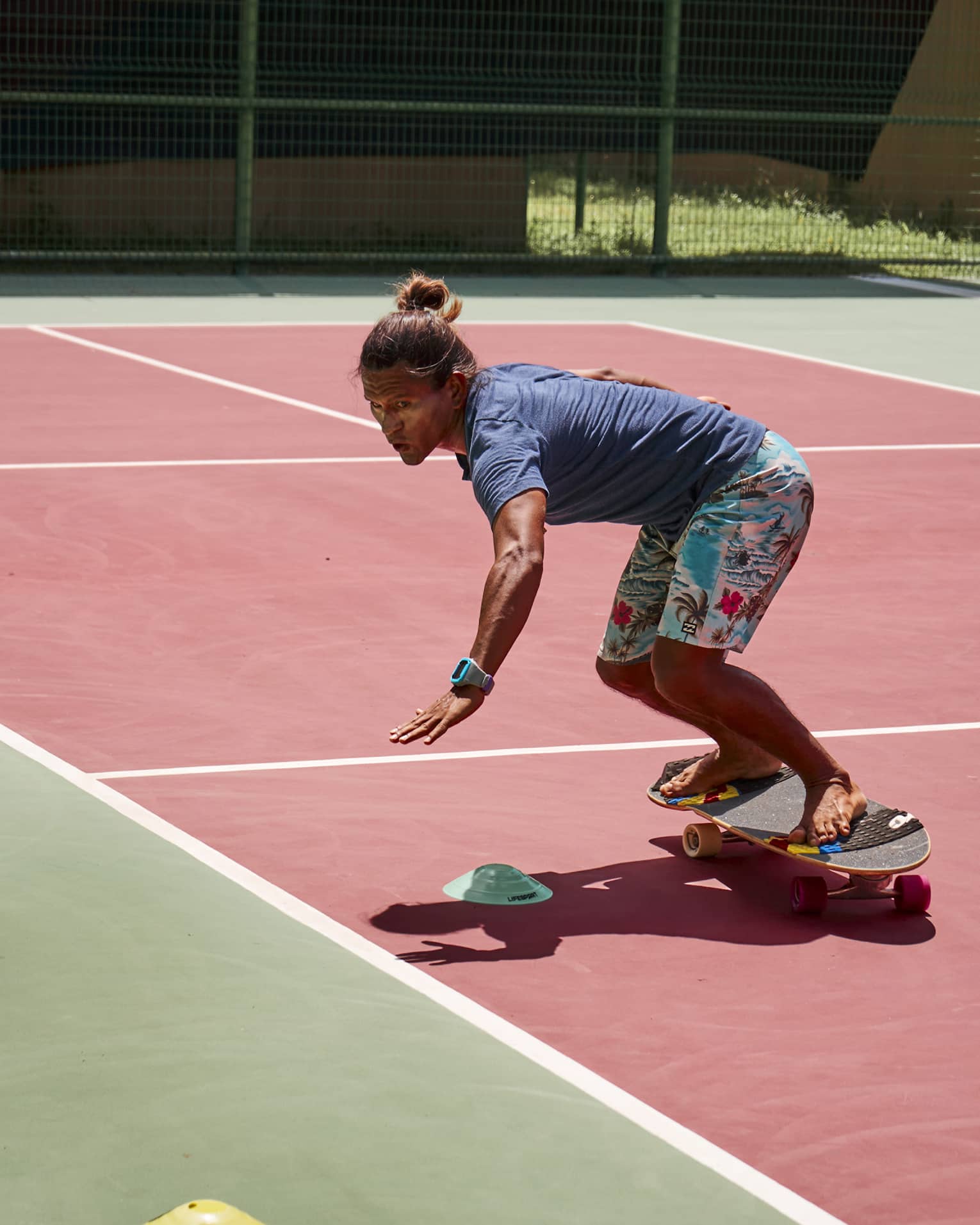 Person wearing light-blue, Hawaiian-print board shorts and a navy t-shirt rides a skateboard around a cone on a tennis court