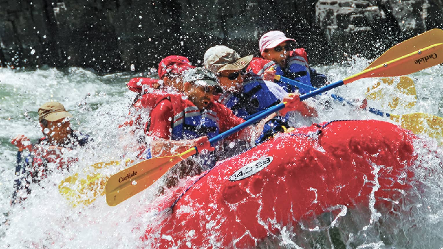 Water sprays around red inflatable raft, people with paddles, life vests white water rafting