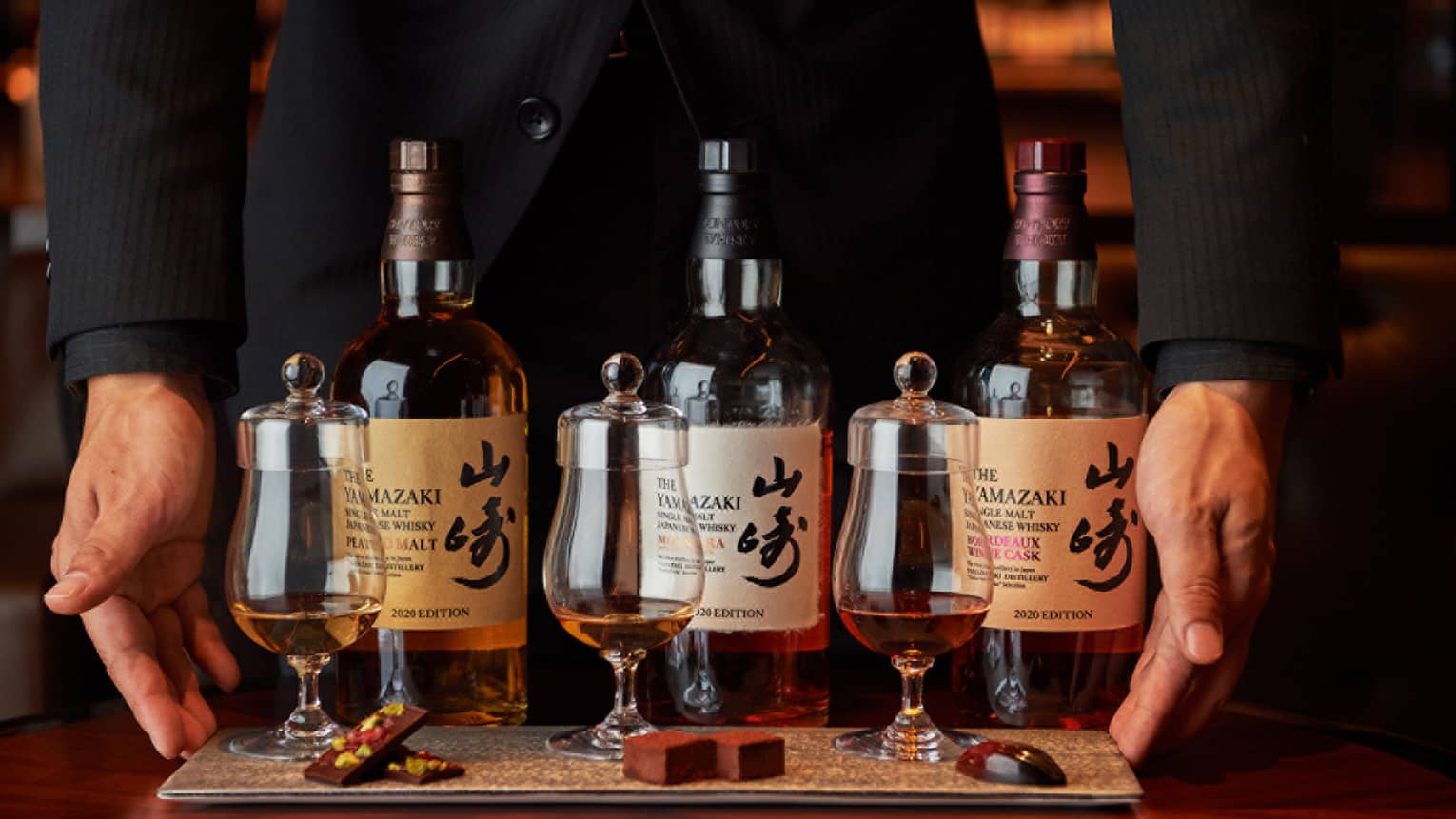 Hands frame a row of three bottles of single malt Japanese whisky behind three curved whisky tumblers with tasting caps.