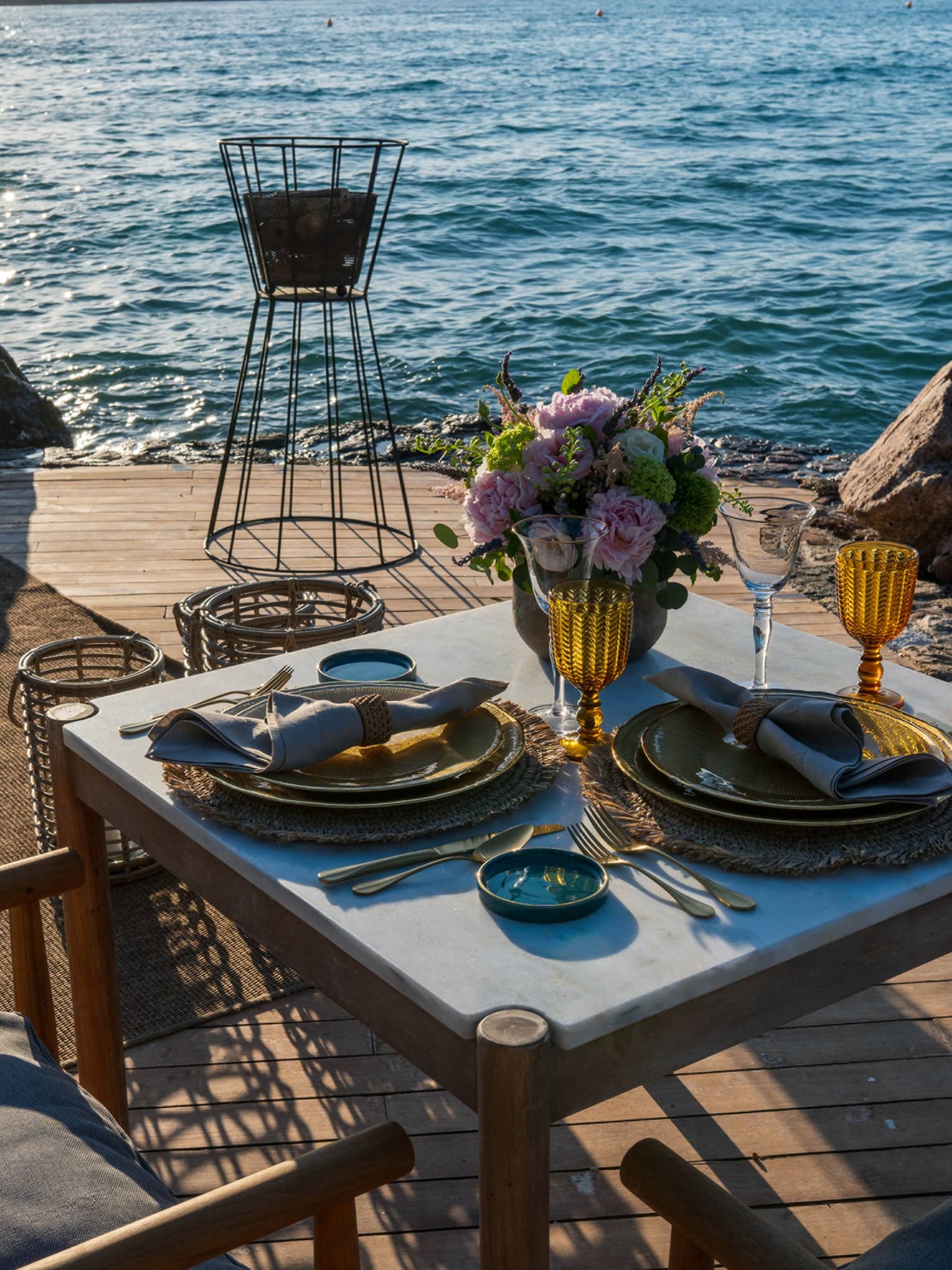 Elegant flowers and two rustic gold place settings with amber wine glasses grace a dining table on a waterfront patio.