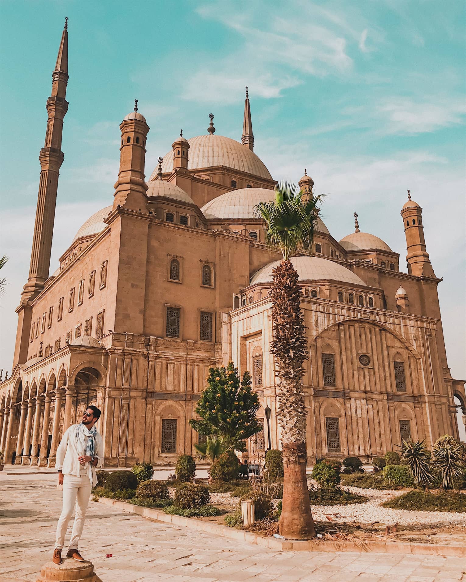 A tourist poses in front of the imposing Mosque of Muhammad Ali, minarets towering above the many domes of the building.