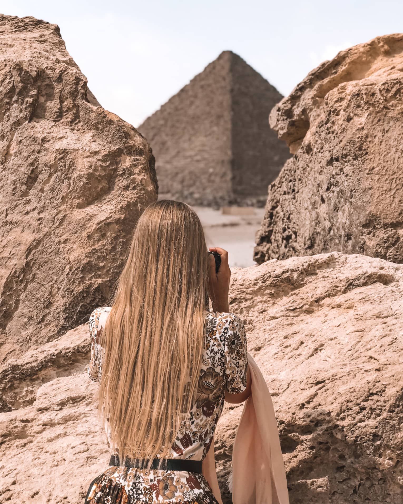 A tourist in a floral dress stands between two large rocks and aims her camera at an enormous pyramid beyond. 