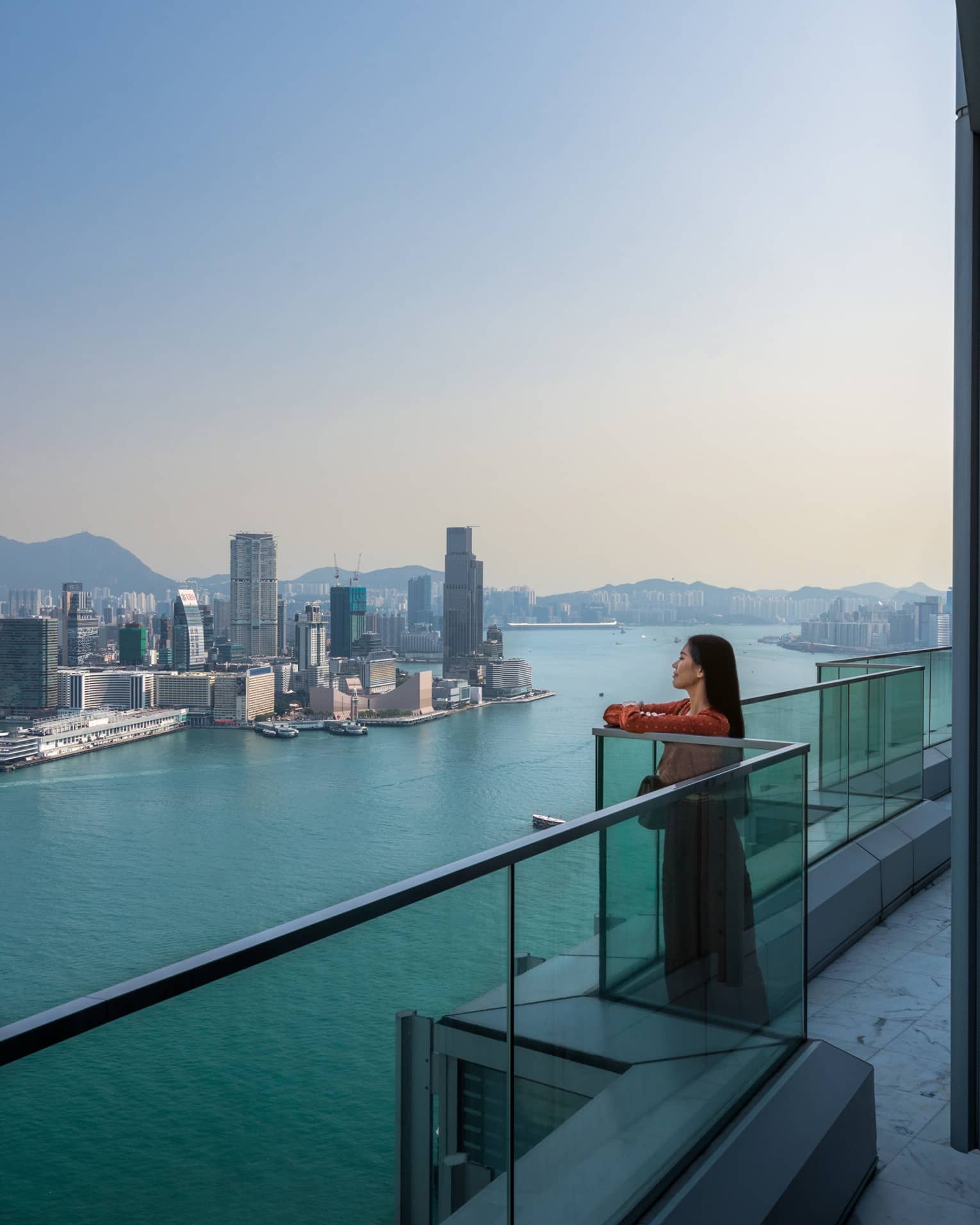 A woman standing on a balcony looking out at water and a city.