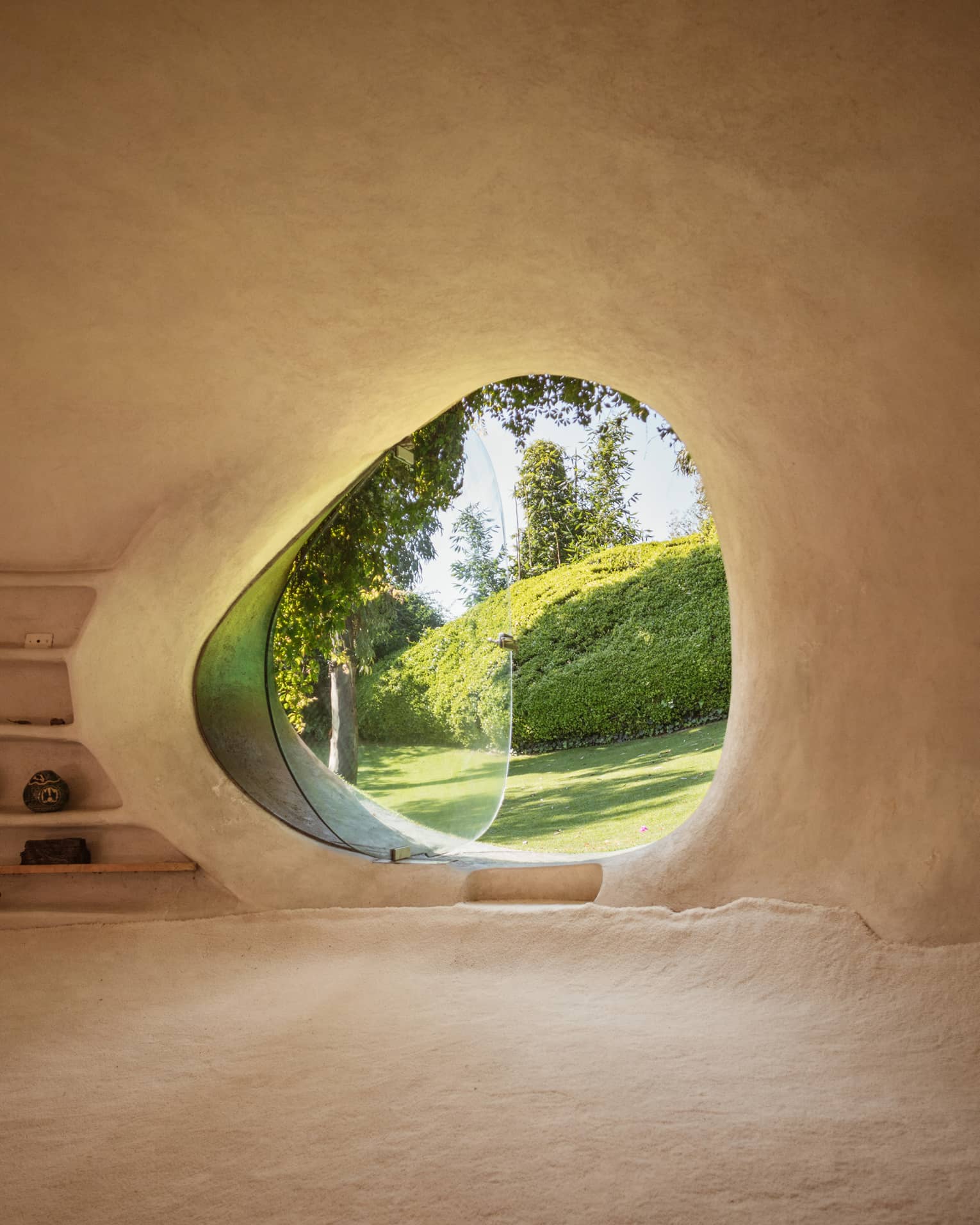 Smooth, adobe wall frames a curved, asymmetrical window, glass pivoted out, a verdant yard with large pruned hedge in view.