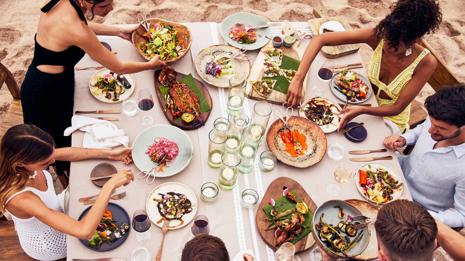 A group of people eating various food on a large table at the beach.