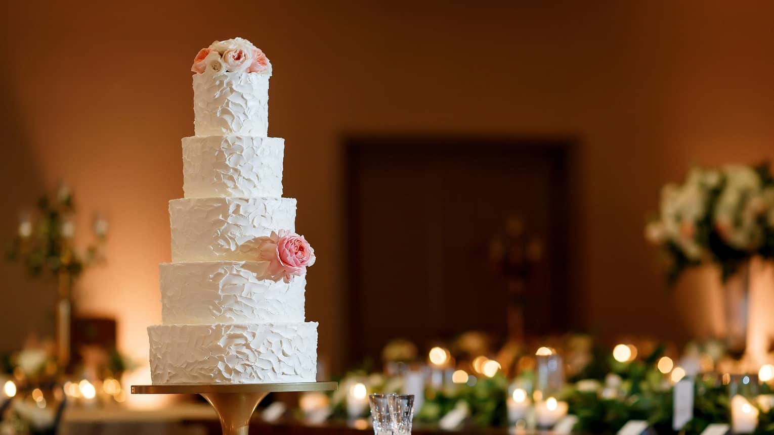 A five-tiered white wedding cake garnished with a white rose and some small white and pink flowers on the top tier , sits on a white round table surrounded by white candles