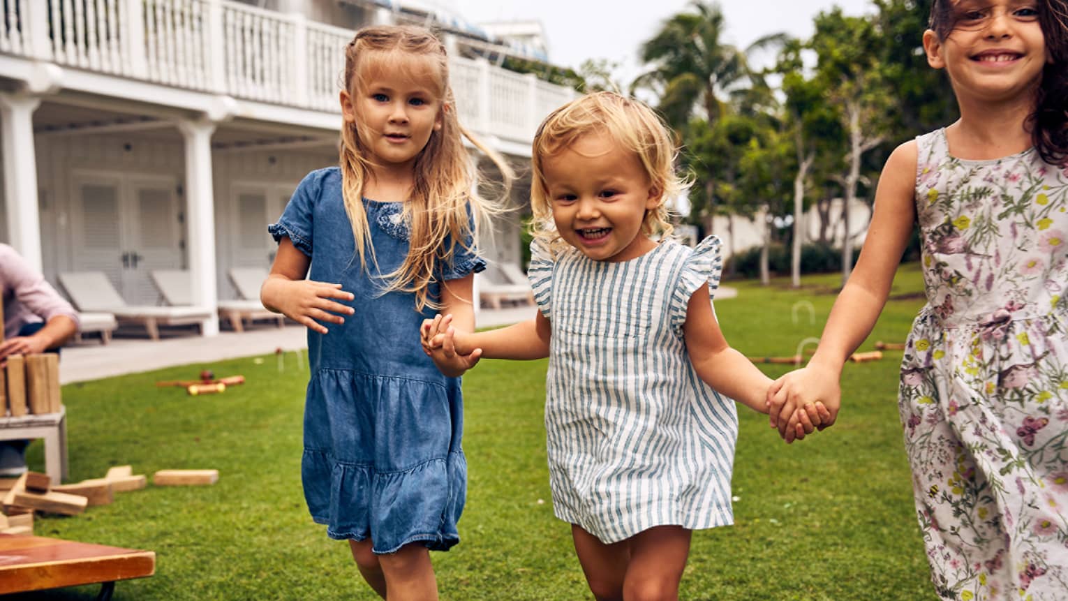 Three young girls holding hands as they play on grass next to a large white building.