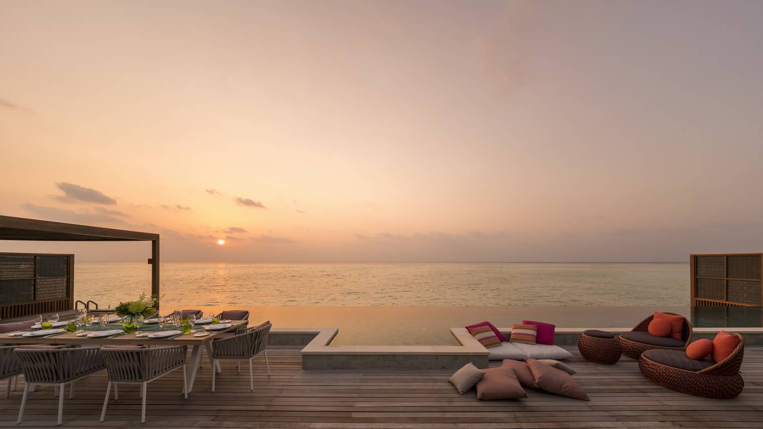Large wooden deck with dining table and lounge chairs, overlooking an ocean sunset