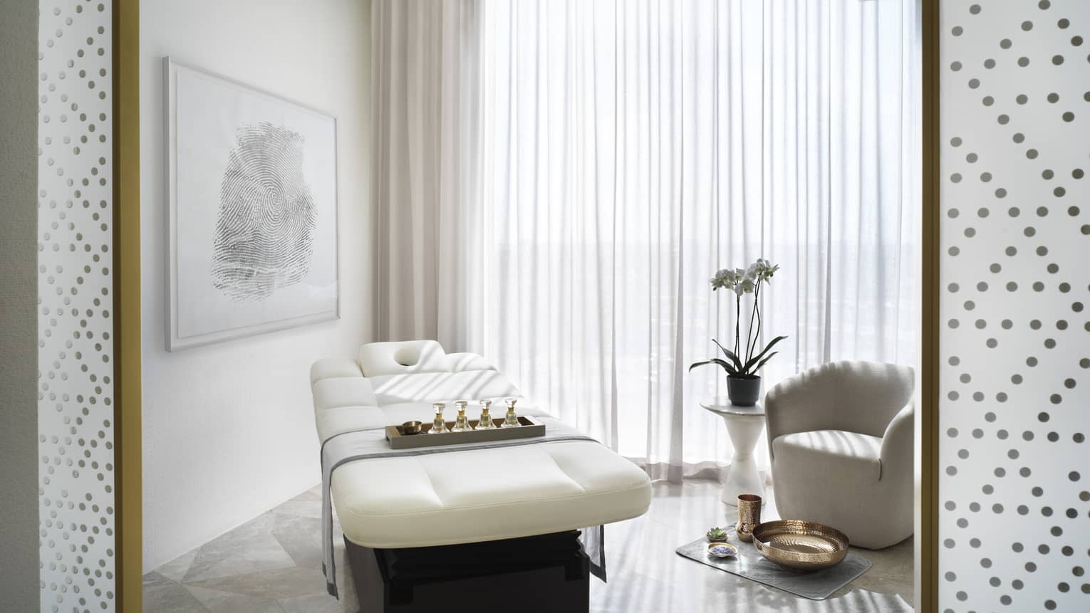 Spa room with a white massage table and white chair, next to a large window covered by a sheer curtain