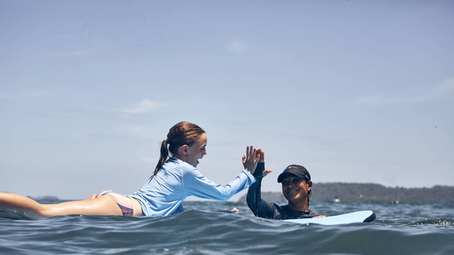 Amid gently rippling waves, a smiling child lies on a surfboard and high-fives an encouraging instructor in the water.