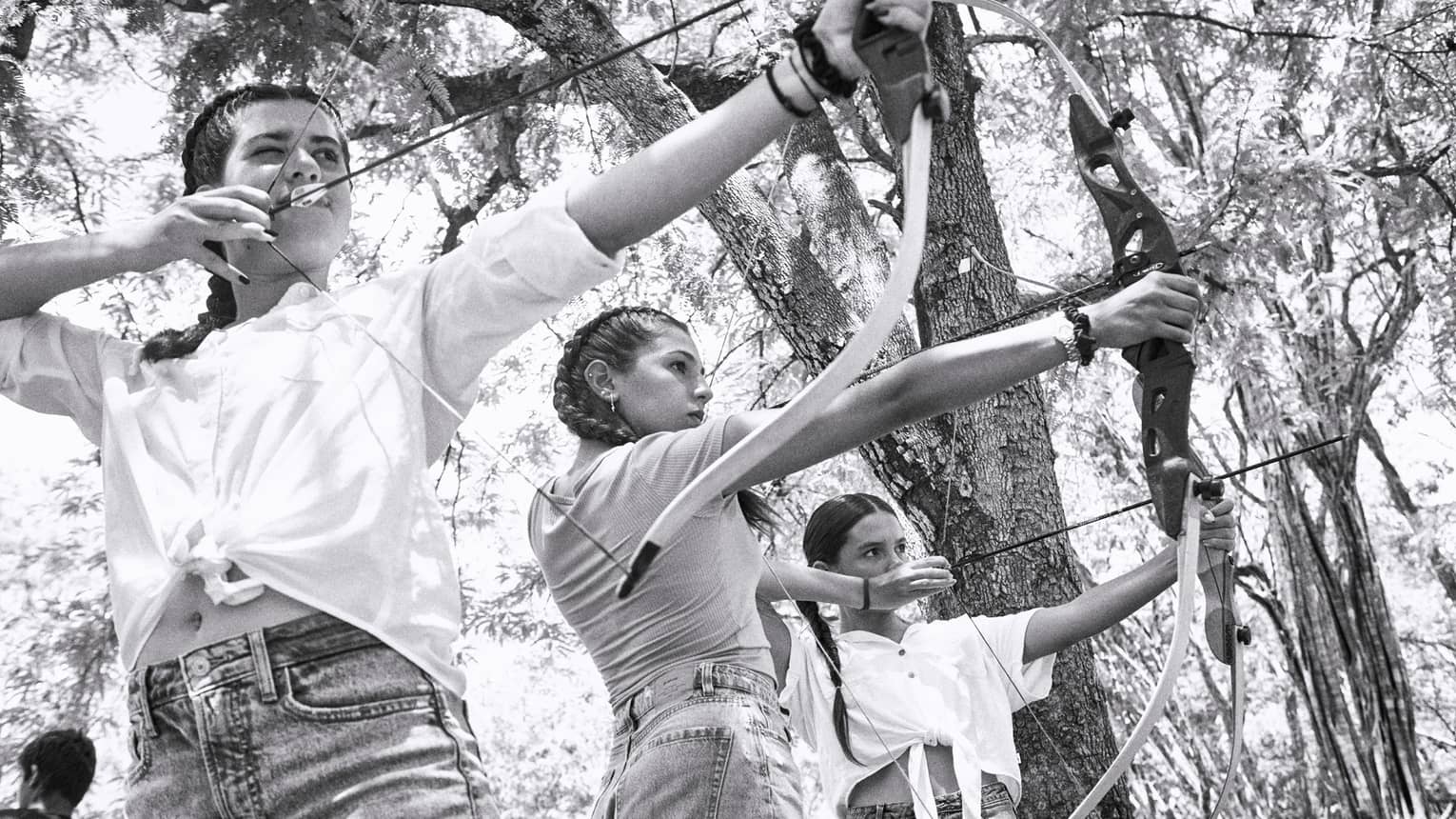 Black and white photo of three young girls aiming traditional archery bows in a forest