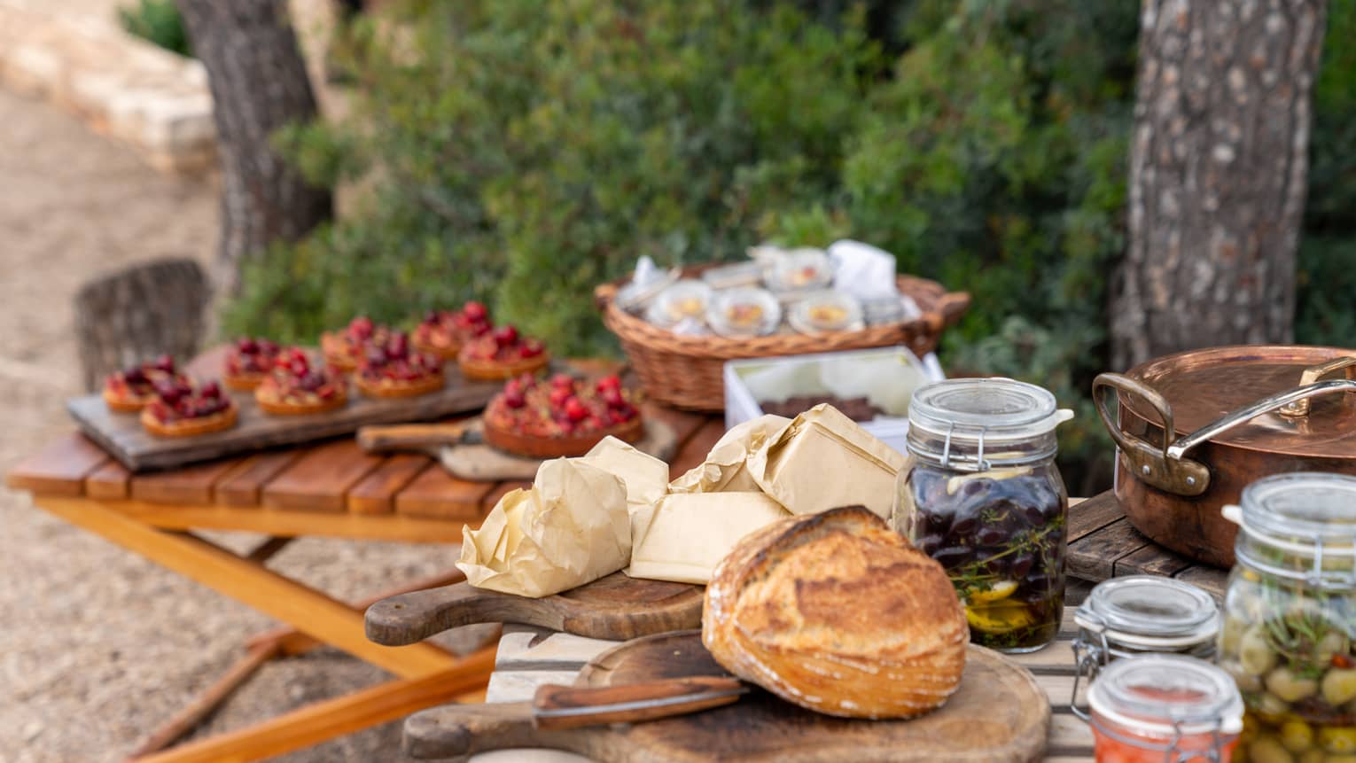 Amid trees and shrubs, a picnic featuring jars of green and black olives, fresh bread over a cutting board and fruit tarts.