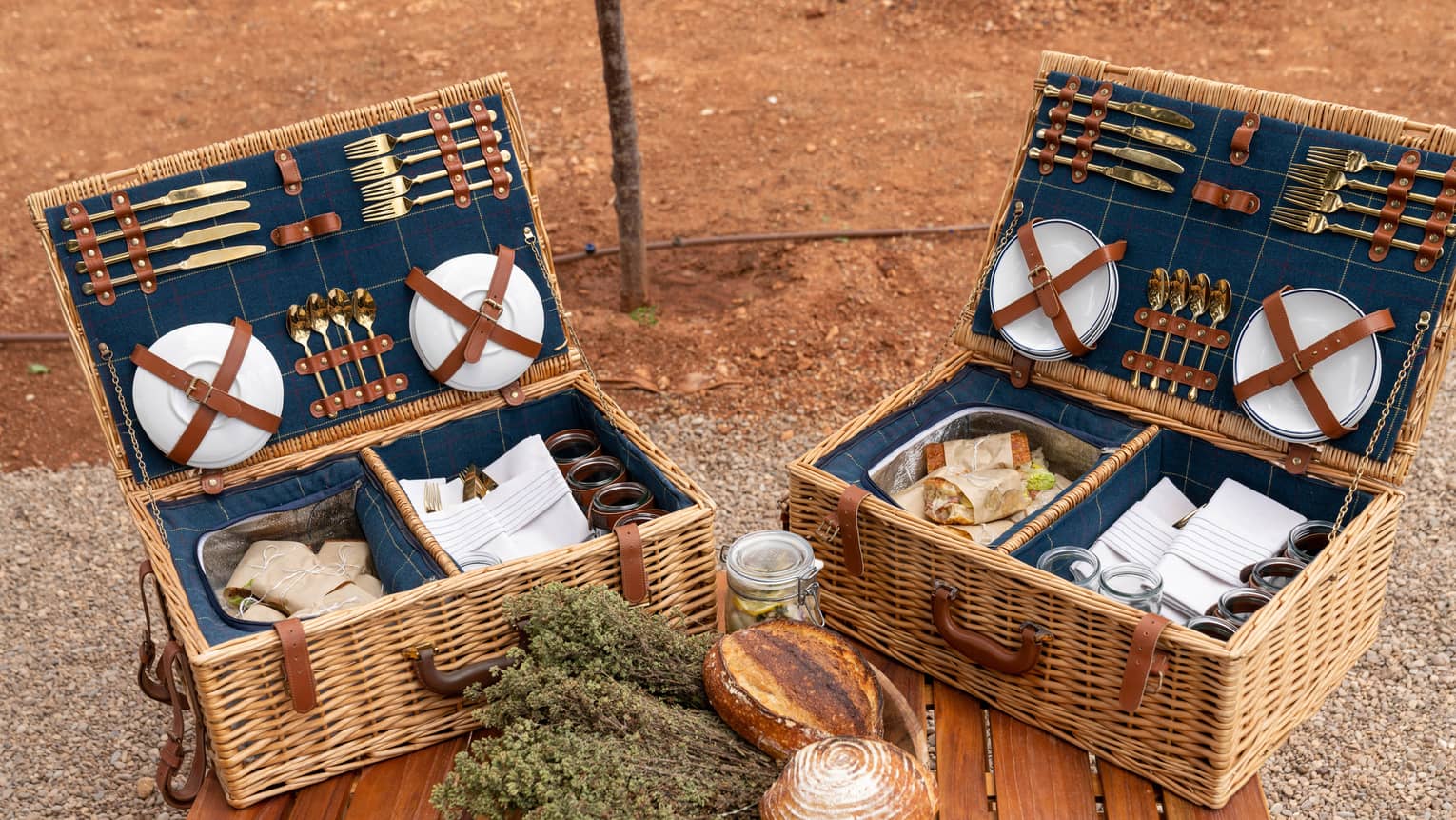 Plates and gold cutlery secured to the lids of picnic baskets containing parchment-wrapped sandwiches, napkins, and glasses.