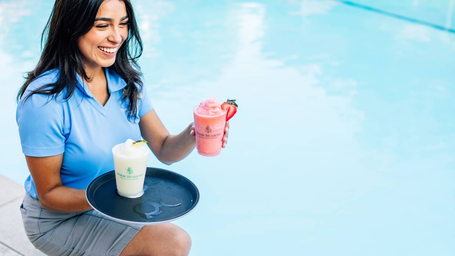Staff member smiling in blue polo shirt kneeling at poolside with tray and two frozen drinks