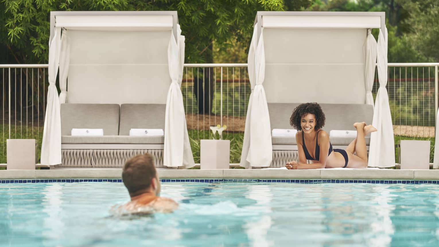 A man swimming in a pool outside with a woman resting under a cabana near the pool.