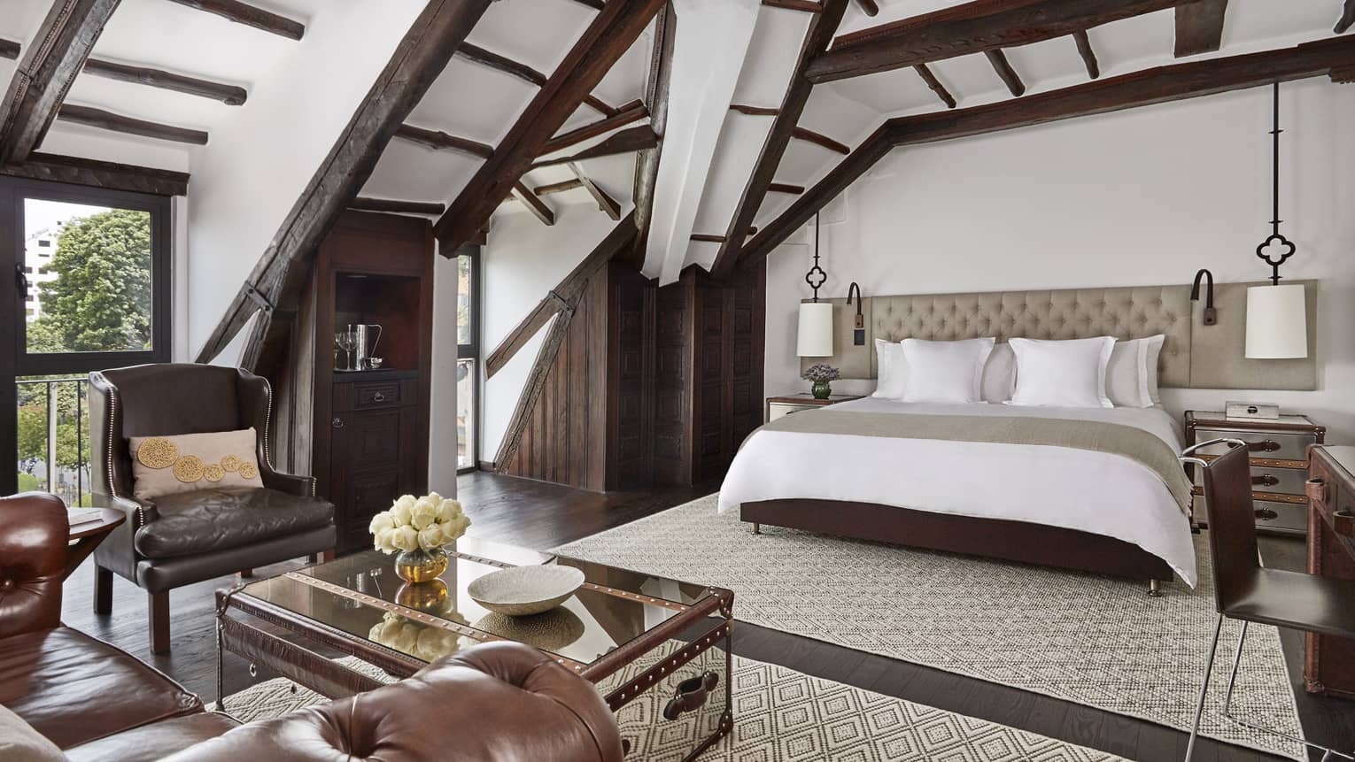 Sloped beam loft ceiling over hotel bed with padded headboard, brown leather armchair, table with flowers