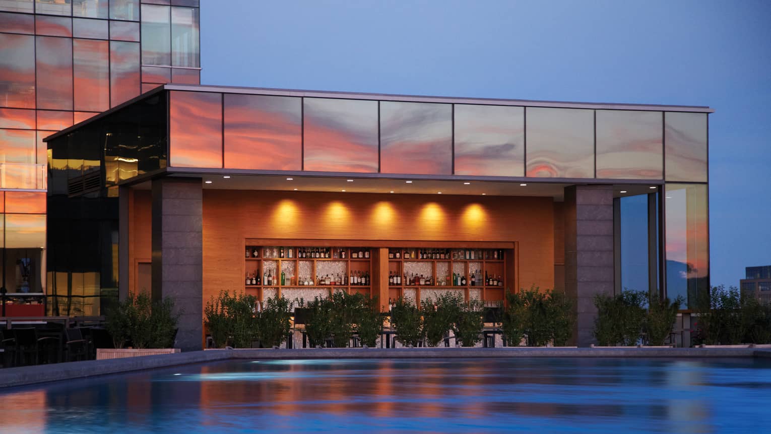 Illuminated swimming pool, wood bar and liquor displays under glass building exterior at sunset