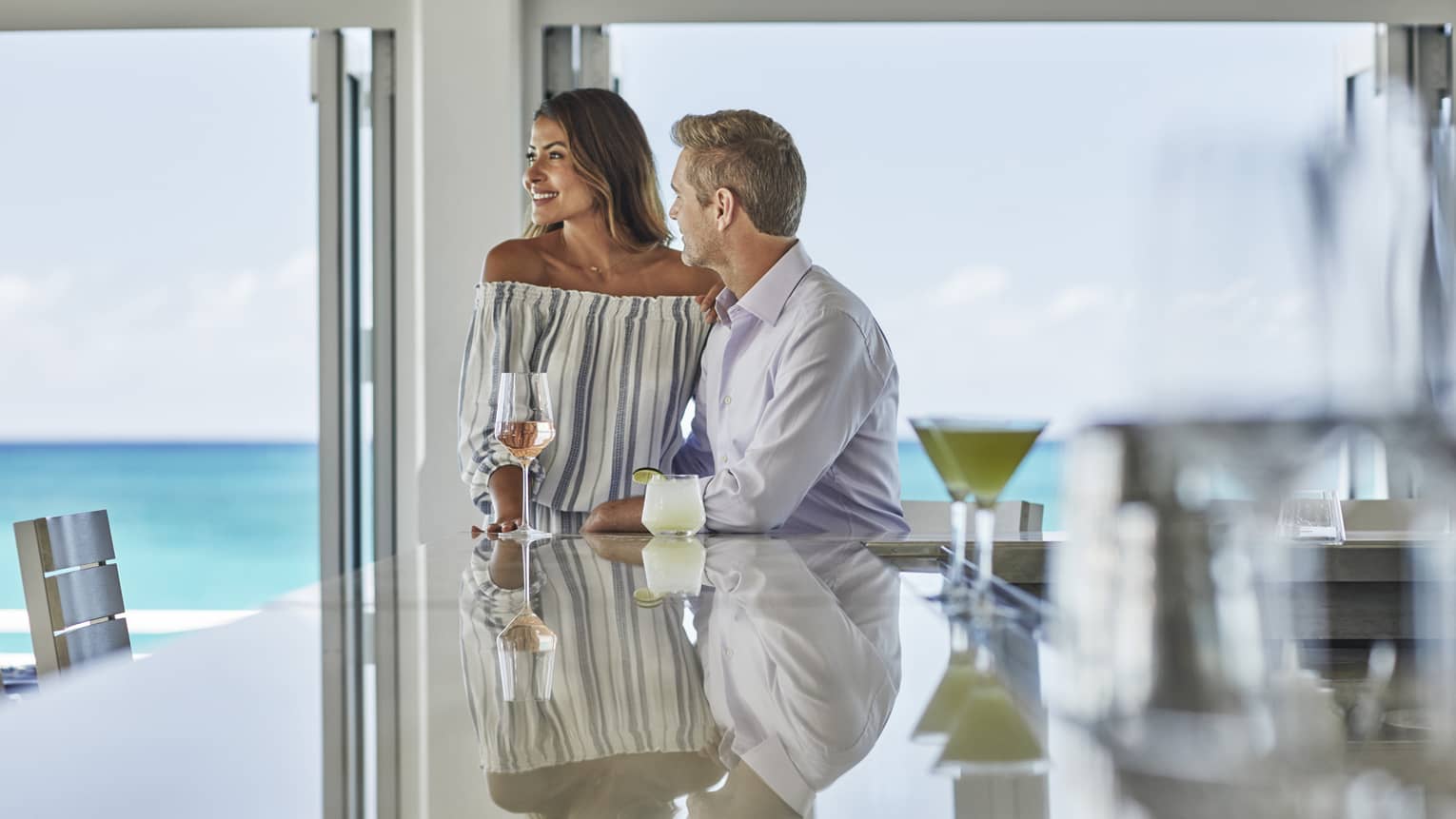Couple with cocktails at end of shining bar by bright windows overlooking ocean