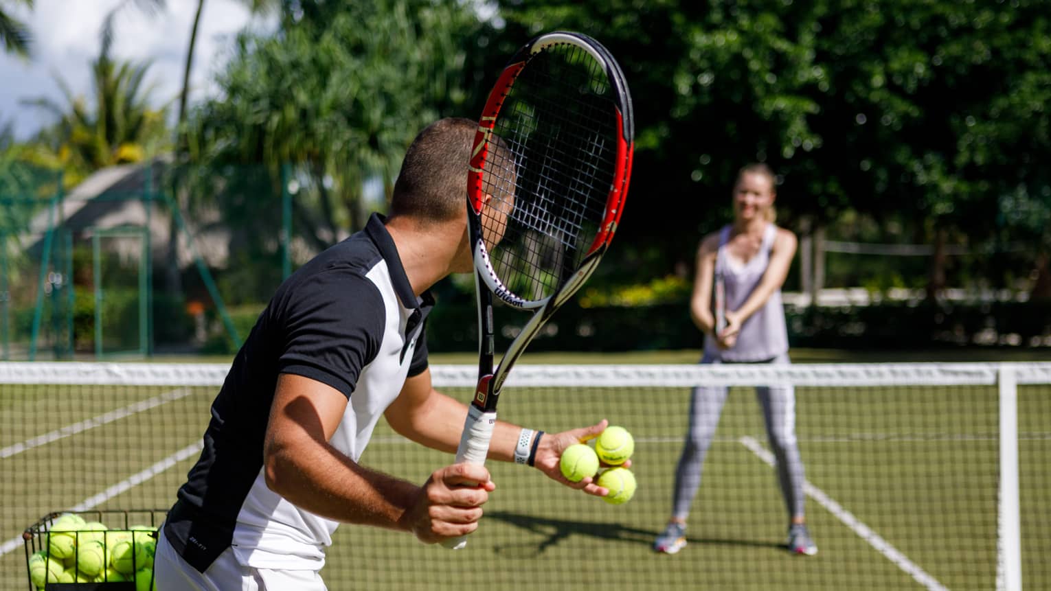 Couple playing tennis on court, man prepares to serve tennis balls to woman ready to swing