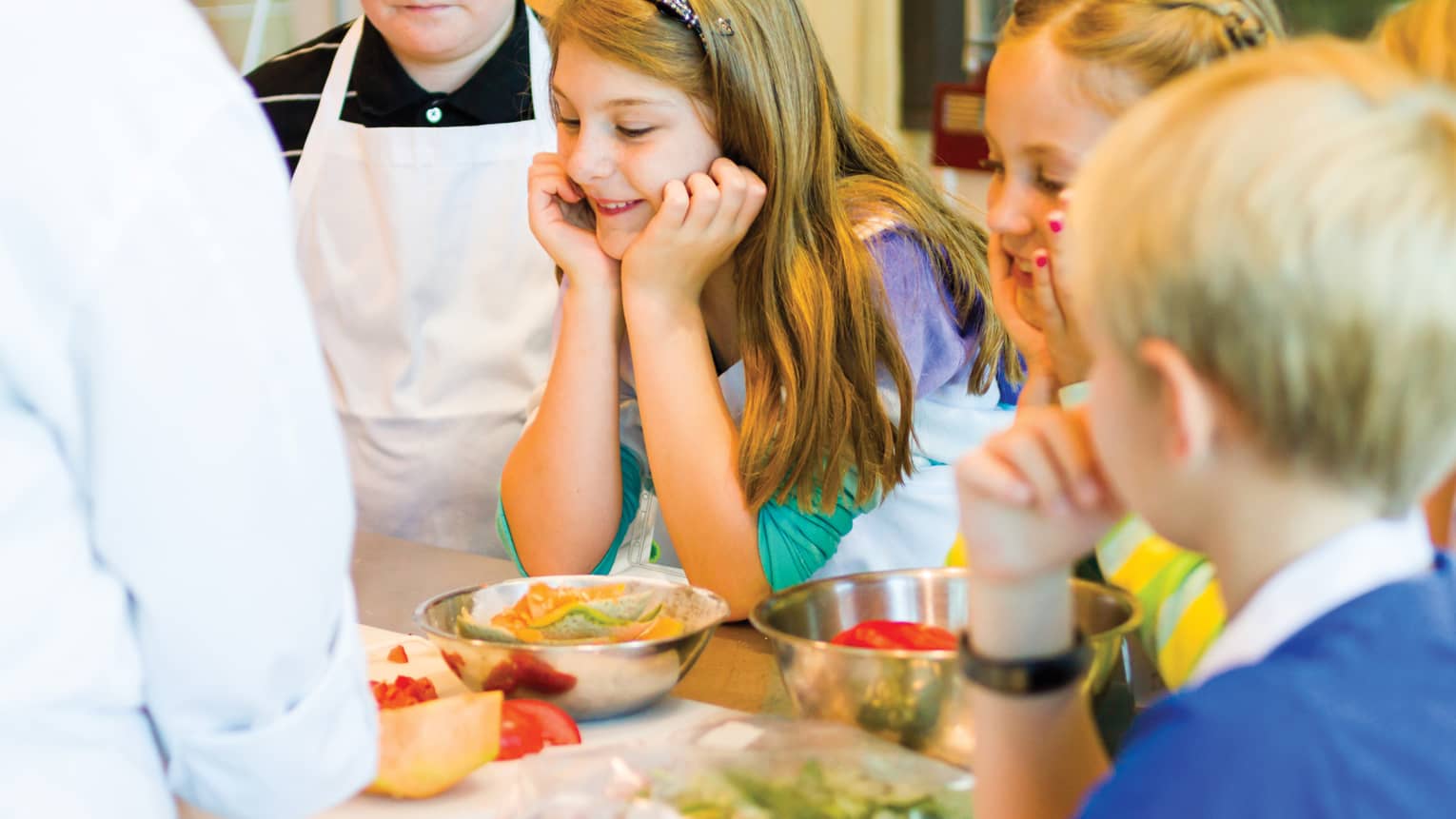 Smiling girl on apron rests chin on hands by other kids during Thai cooking class