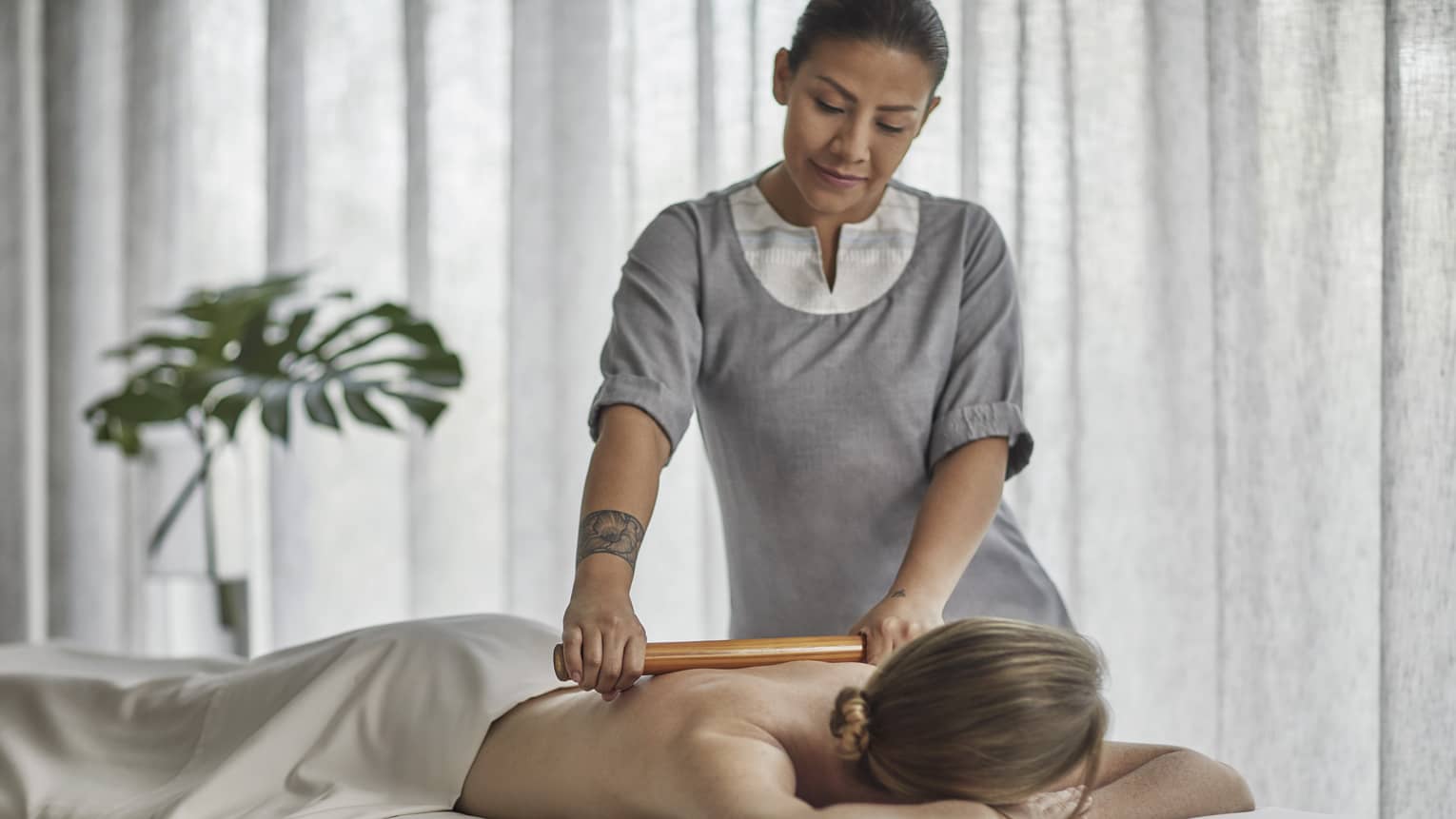 Spa therapist wearing a grey tunic rolls a wooden roller on the bare back of a guest who is laying on a massage table 