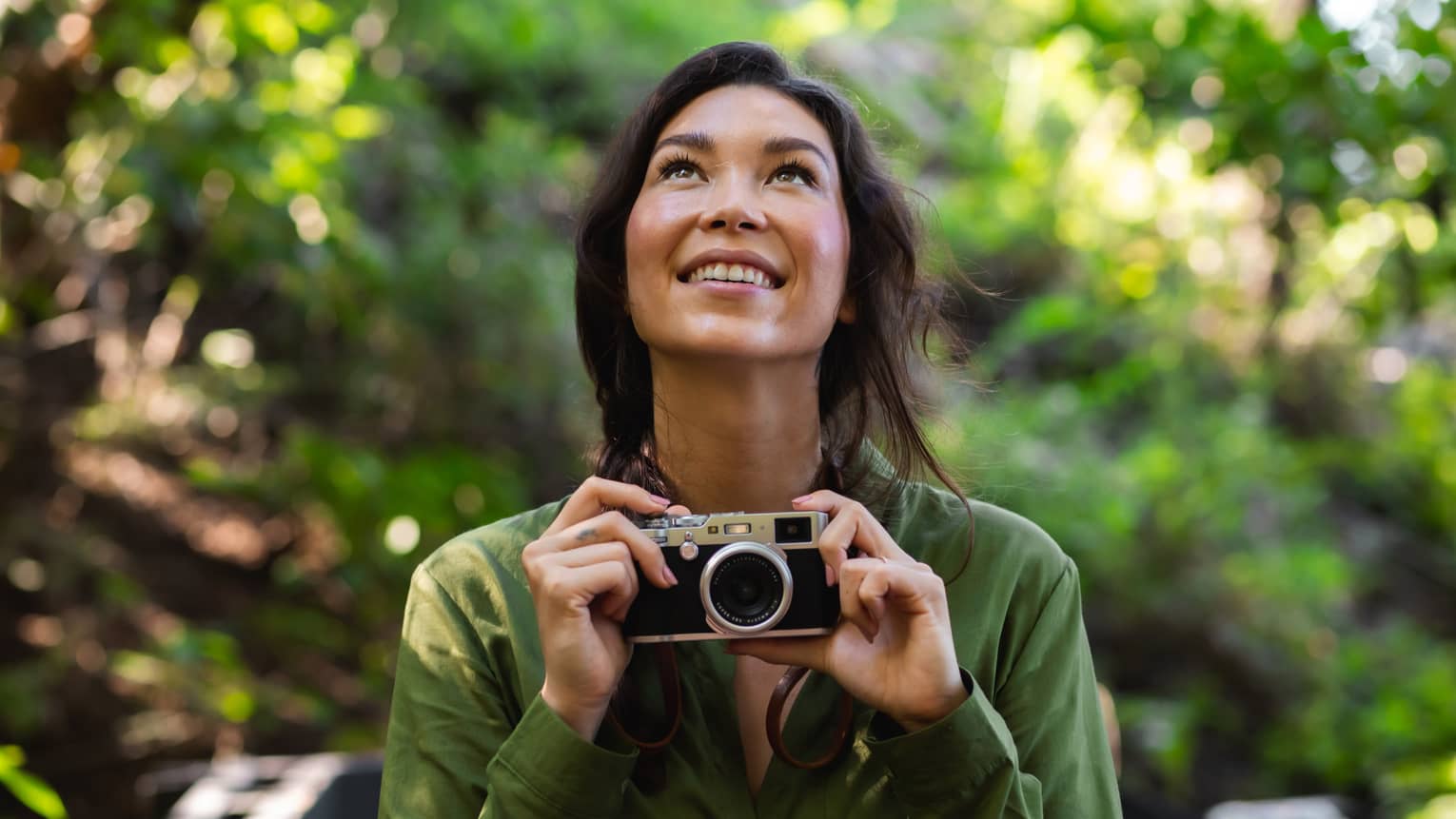 Woman with long dark hair wearing a green button-down shirt holds a camera as she looks upward in the forest