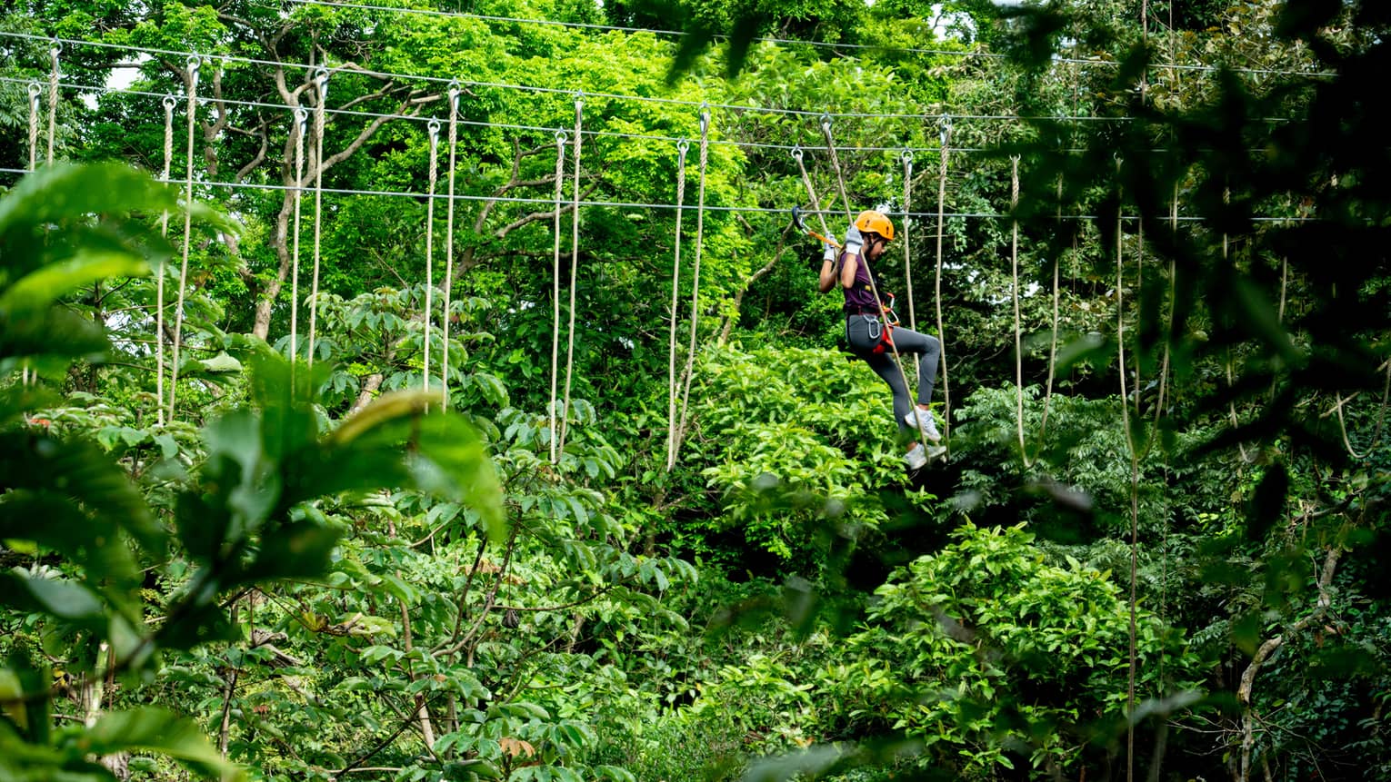 A person wearing a yellow helmet and dark clothing swings on an aerial ropes course set in the lush green canopy of the Costa Rica rainforest