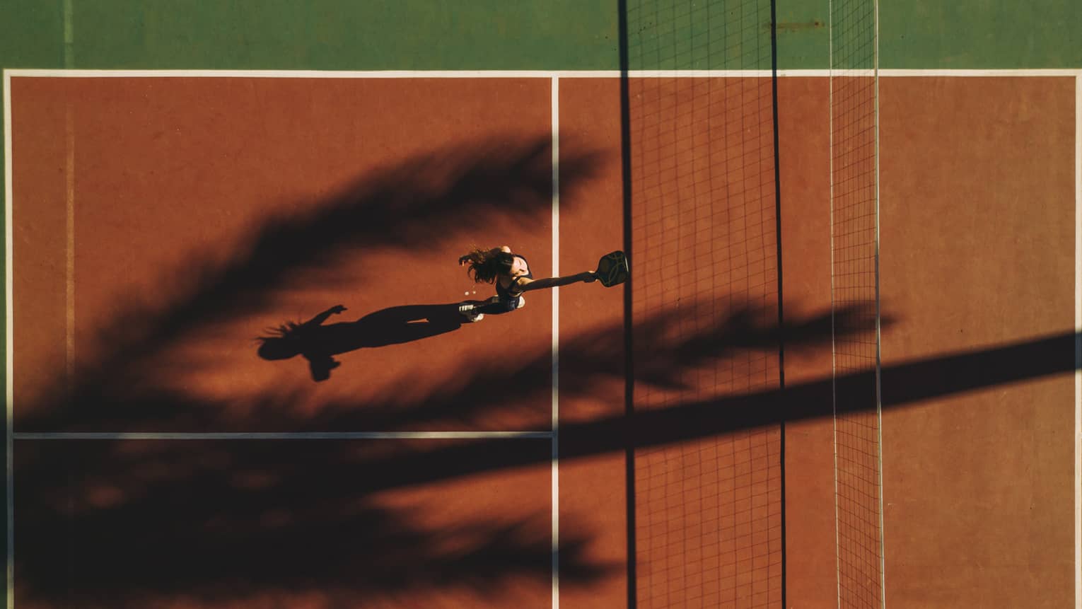 A palm tree casts a shadow on a terracotta-coloured pickleball court as a player stands behind the kitchen, paddle up high.