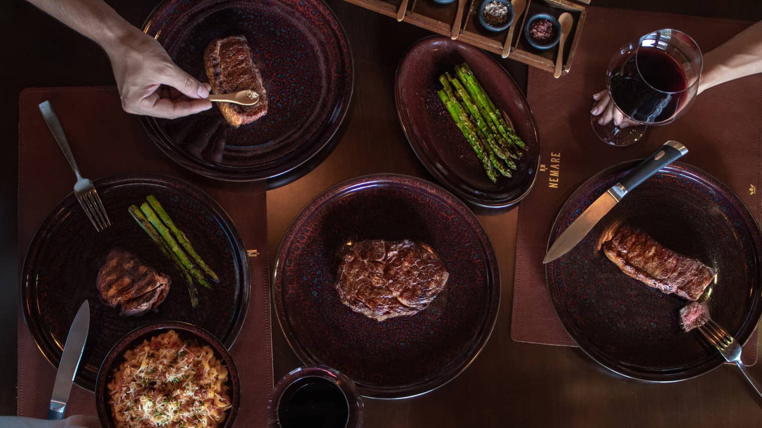 Overhead view of six dark-red stoneware dishes on a wooden table, four each plated with different cuts of steak, one with asparagus and another with a past-type food being passed by a hand