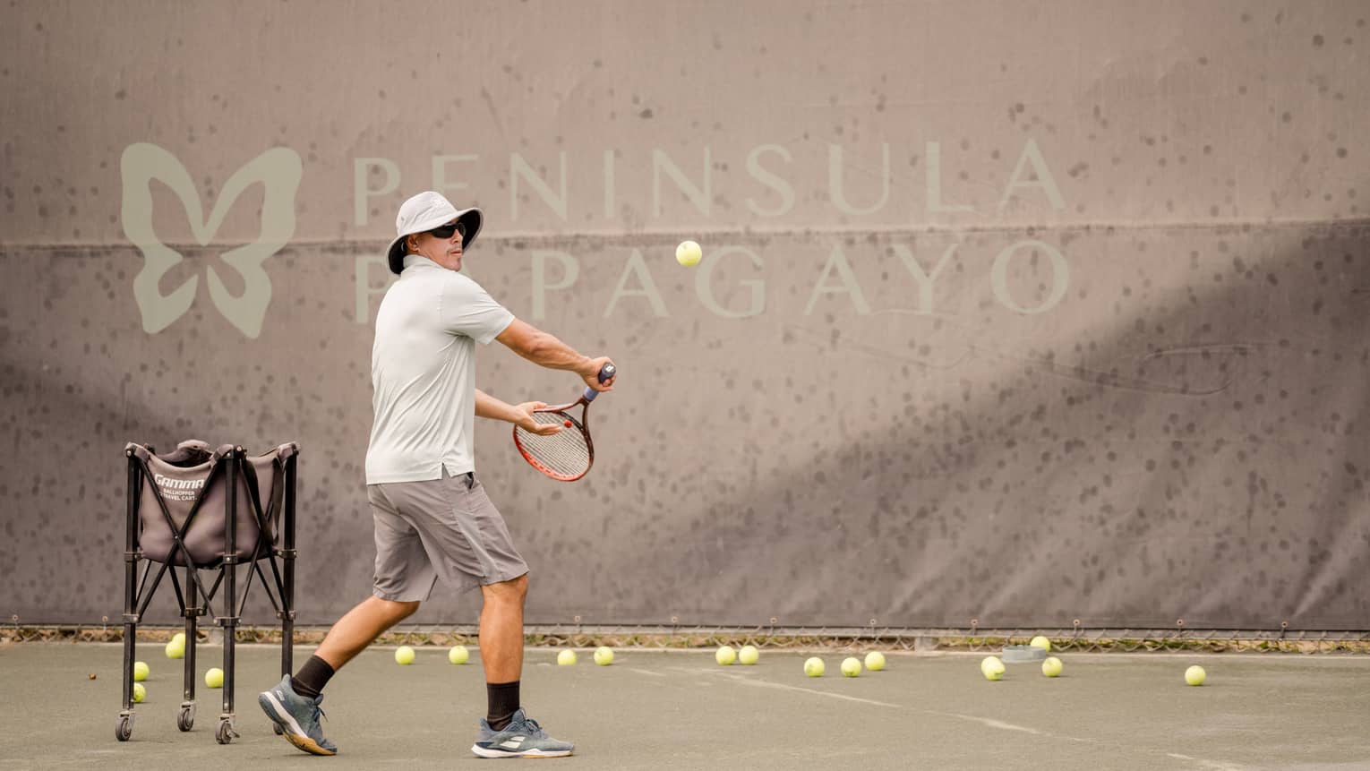 Standing on a clay tennis court beside a ball hopper, balls scattered across the ground, a player prepares to hit a backhand.